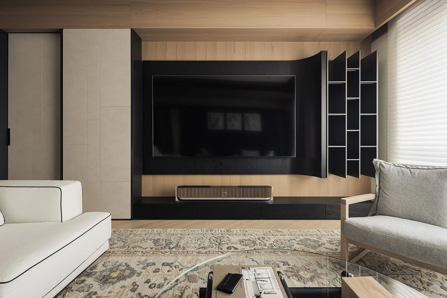 Contemporary living room featuring a large curved black media wall with built-in TV and open shelving, light wood paneling, beige sofa, grey armchair, and patterned area rug.