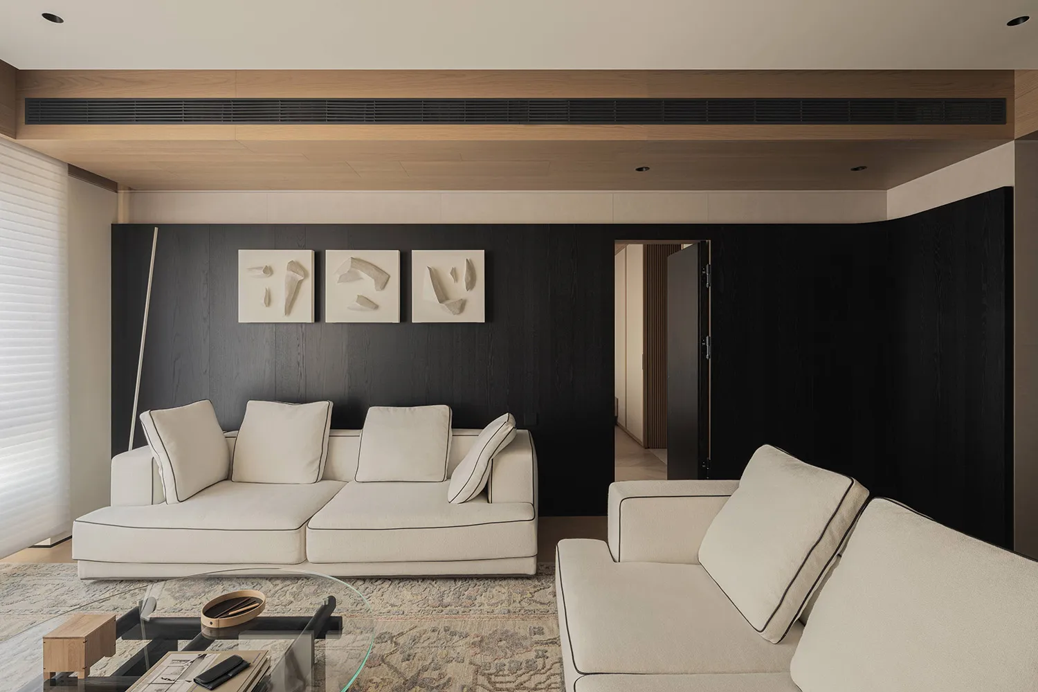 Modern living room featuring a dark wood accent wall with three white abstract artworks, large off-white L-shaped sectional sofa, glass coffee table on a patterned area rug, and wooden ceiling detail.