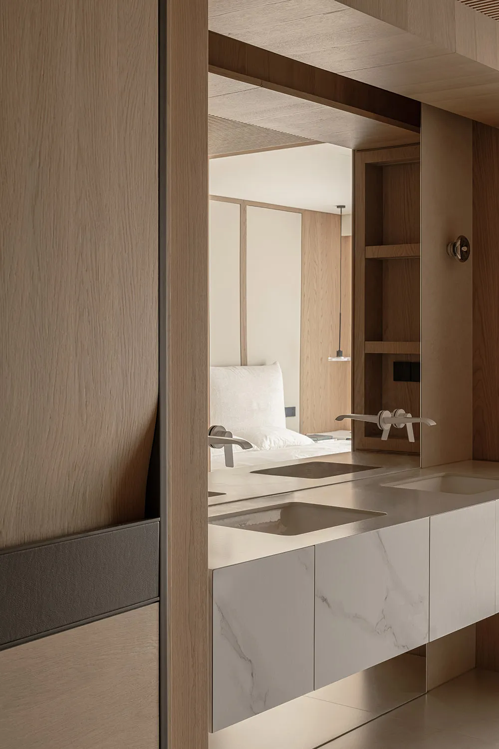 Modern minimalist bathroom with light oak cabinetry, white marble floating double vanity with two integrated sinks, and a large mirror reflecting a bedroom with white bedding.