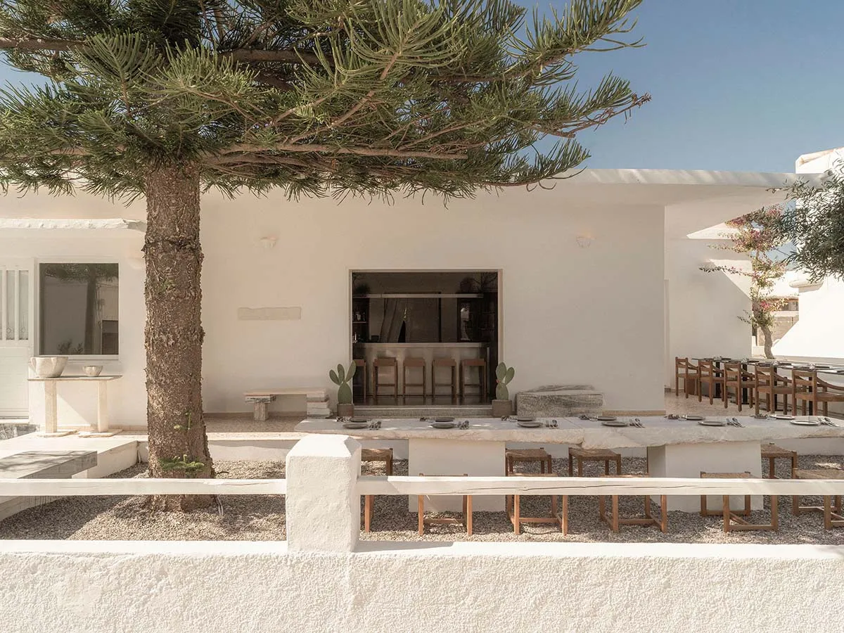 Outdoor dining courtyard with long stone table, wooden stools, whitewashed building, and a large pine tree under clear blue sky