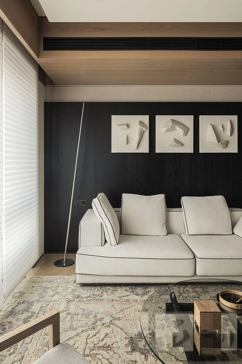 Modern living room featuring a white sectional sofa, dark wood accent wall with three framed sculptural artworks, patterned area rug, and wooden ceiling details.