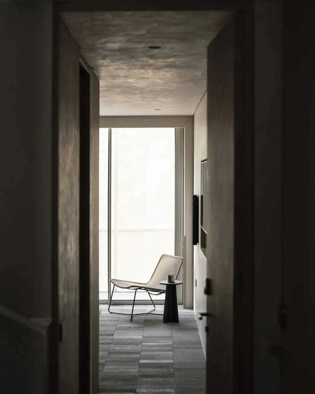 View through a doorway into a minimalist concrete hallway featuring a woven lounge chair, black side table, and large window with natural light.