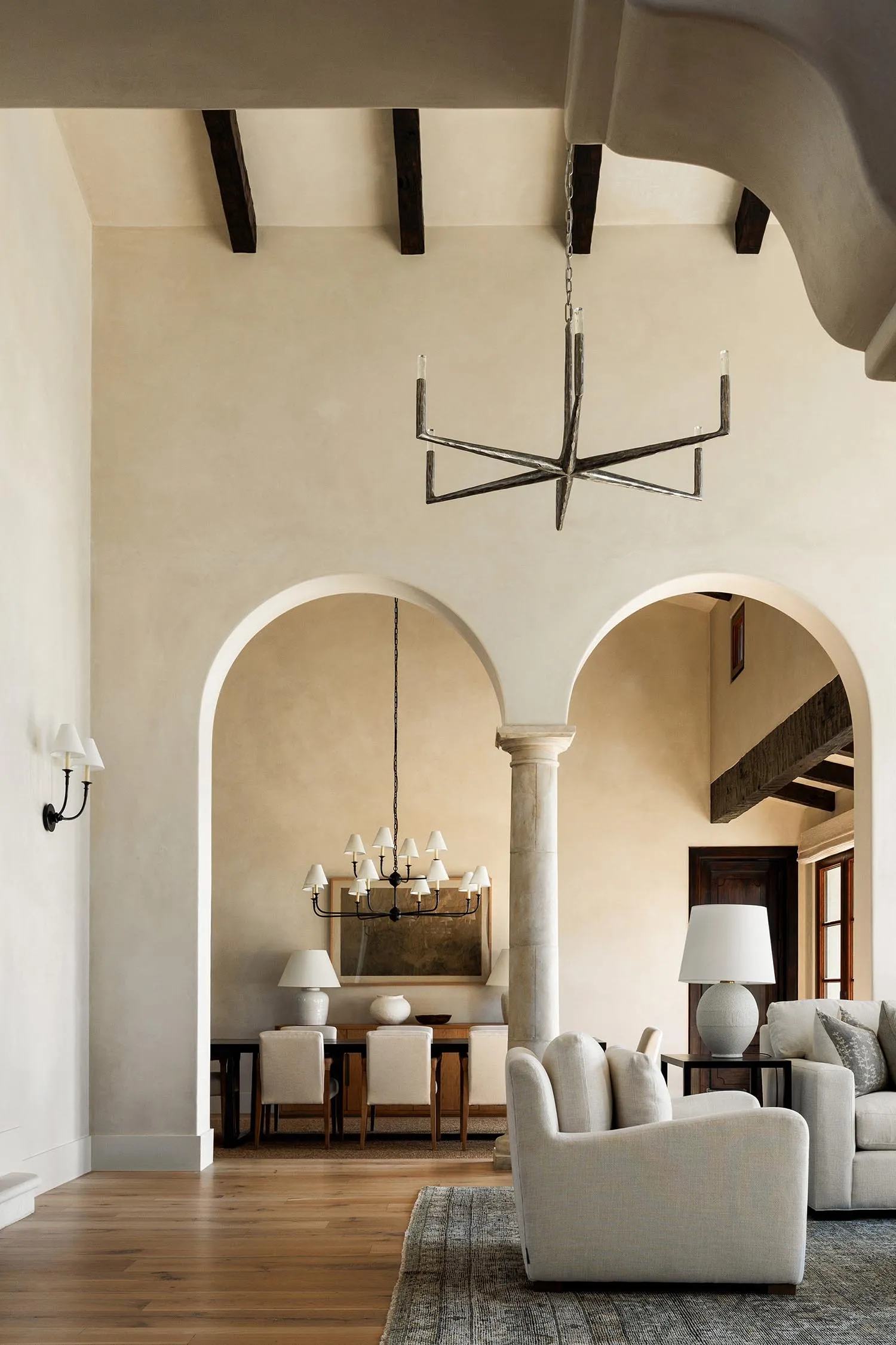 Spacious living and dining area in a Tuscan-style villa featuring high vaulted ceilings with dark wood beams, creamy plaster walls, two large arched openings, a modern starburst iron chandelier, and neutral upholstered furniture.