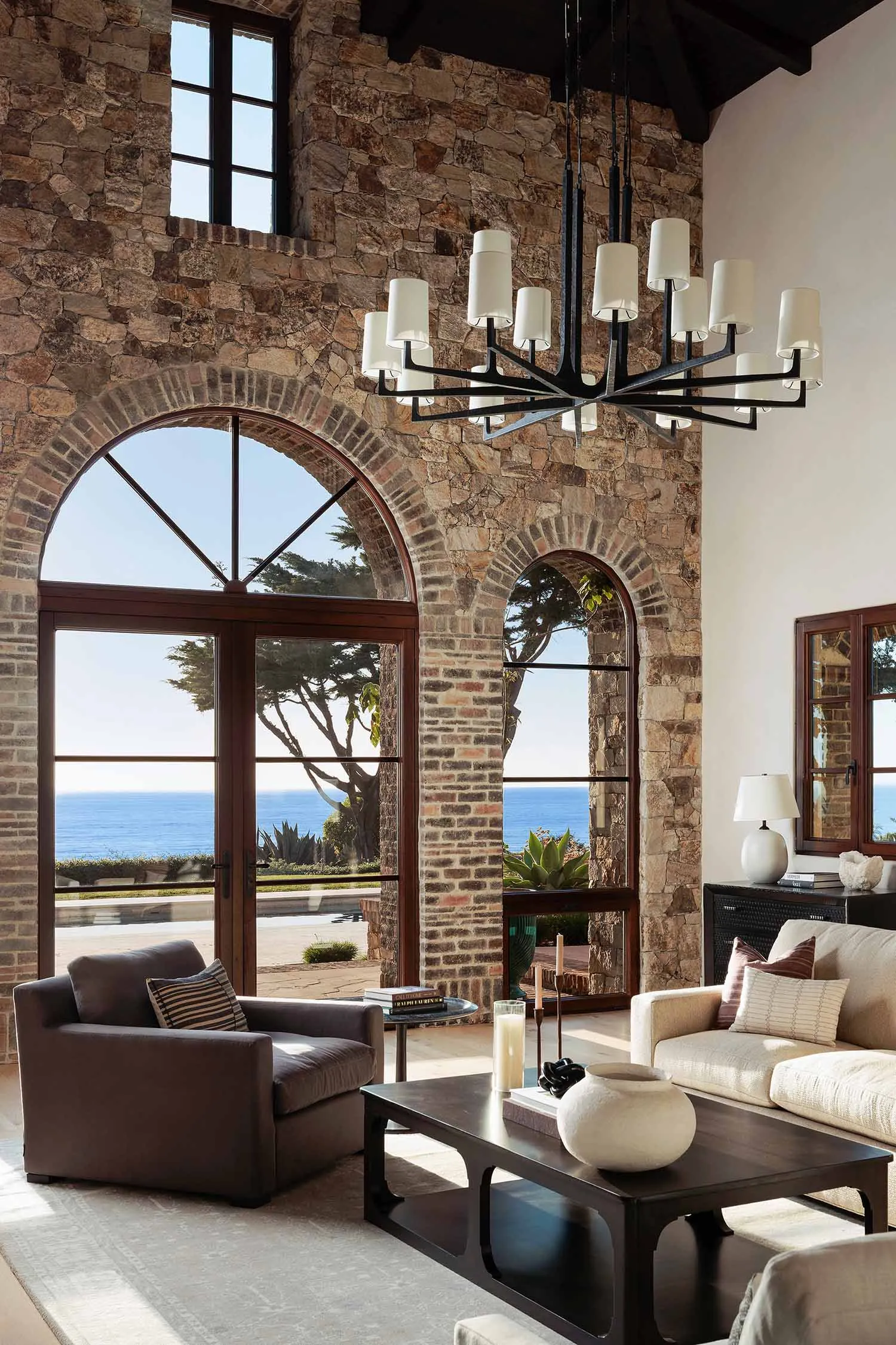Luxurious living room featuring a massive natural stone wall, large arched wooden doors and windows overlooking the ocean, a modern black chandelier, dark leather armchair, and light neutral sofa.