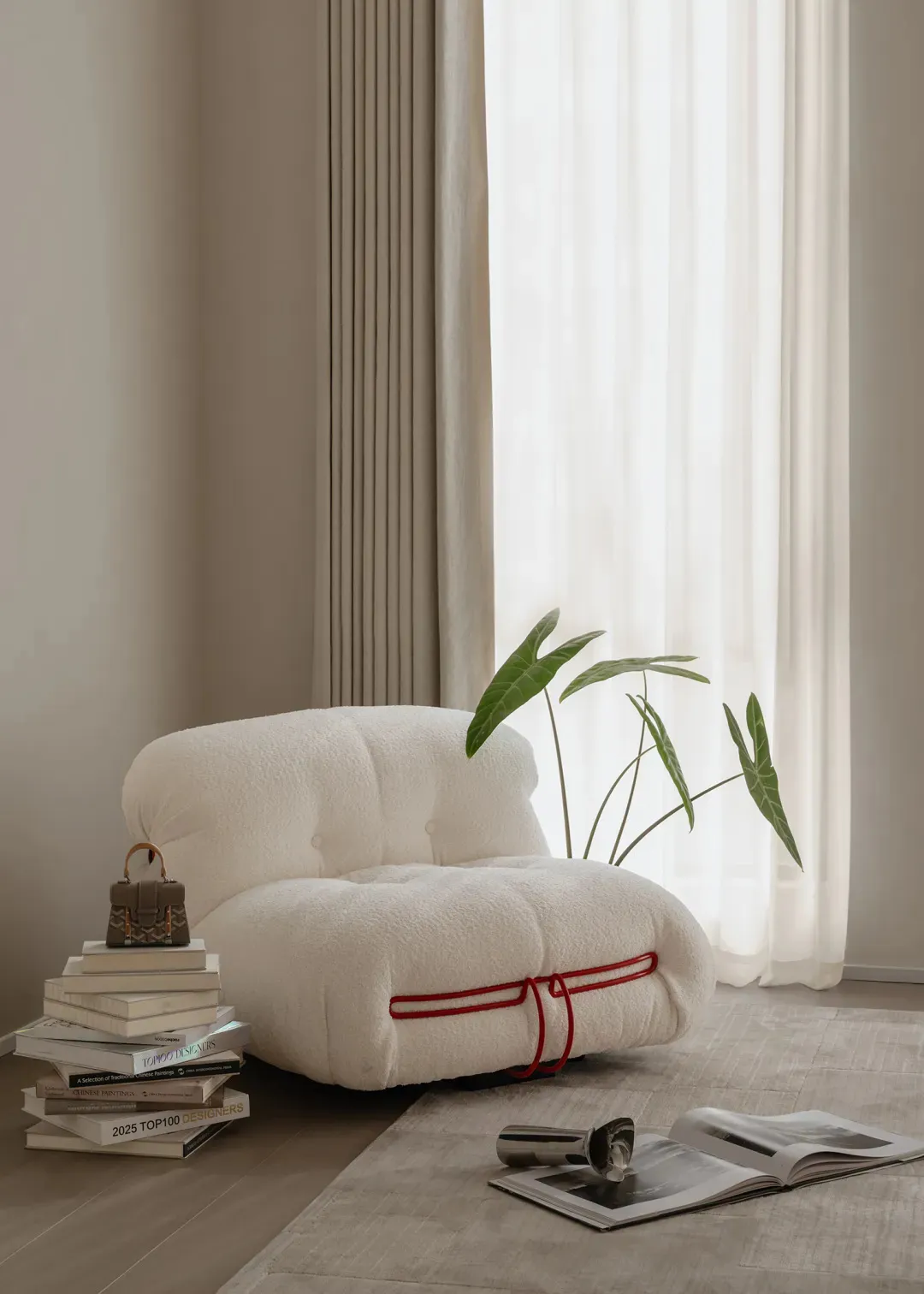 White boucle teddy-bear style lounge chair with red rope detail, positioned next to a stack of design books, a small handbag, an open magazine, and a potted plant in front of a bright window with sheer curtains.