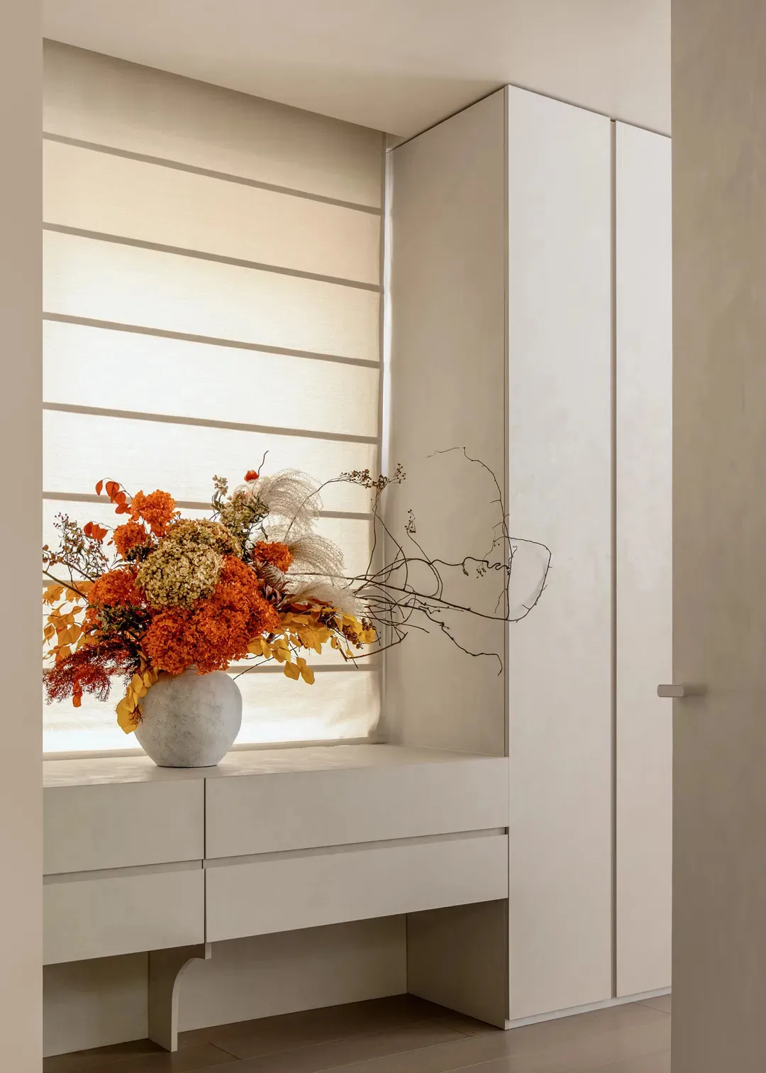 Built-in white storage unit beneath a window with horizontal blinds, featuring a large round marble vase filled with vibrant orange, rust, and cream dried flowers and branches.