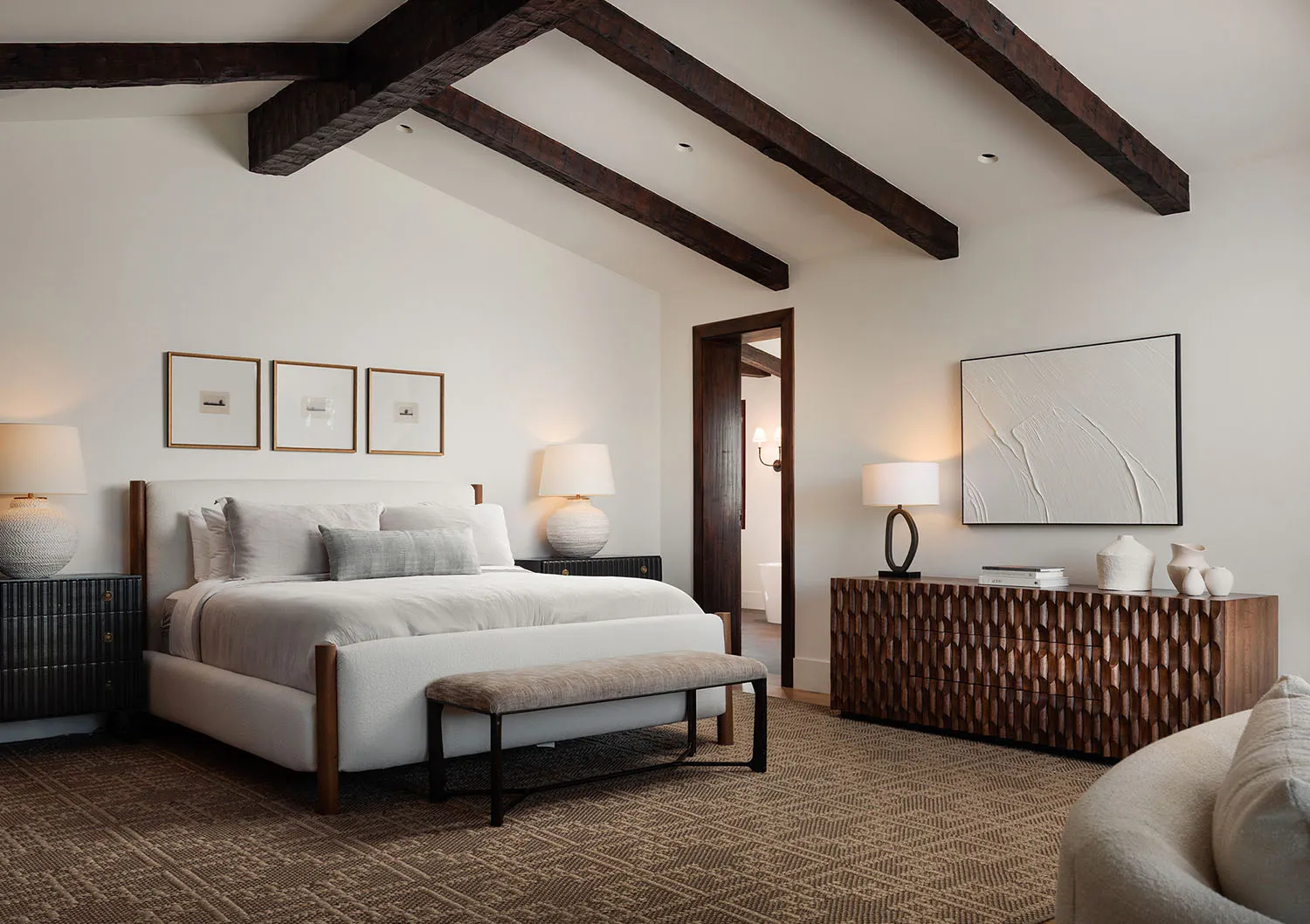 Modern rustic bedroom with white walls, exposed dark wooden ceiling beams, upholstered bed with neutral linens, textured wooden console, and herringbone carpet.