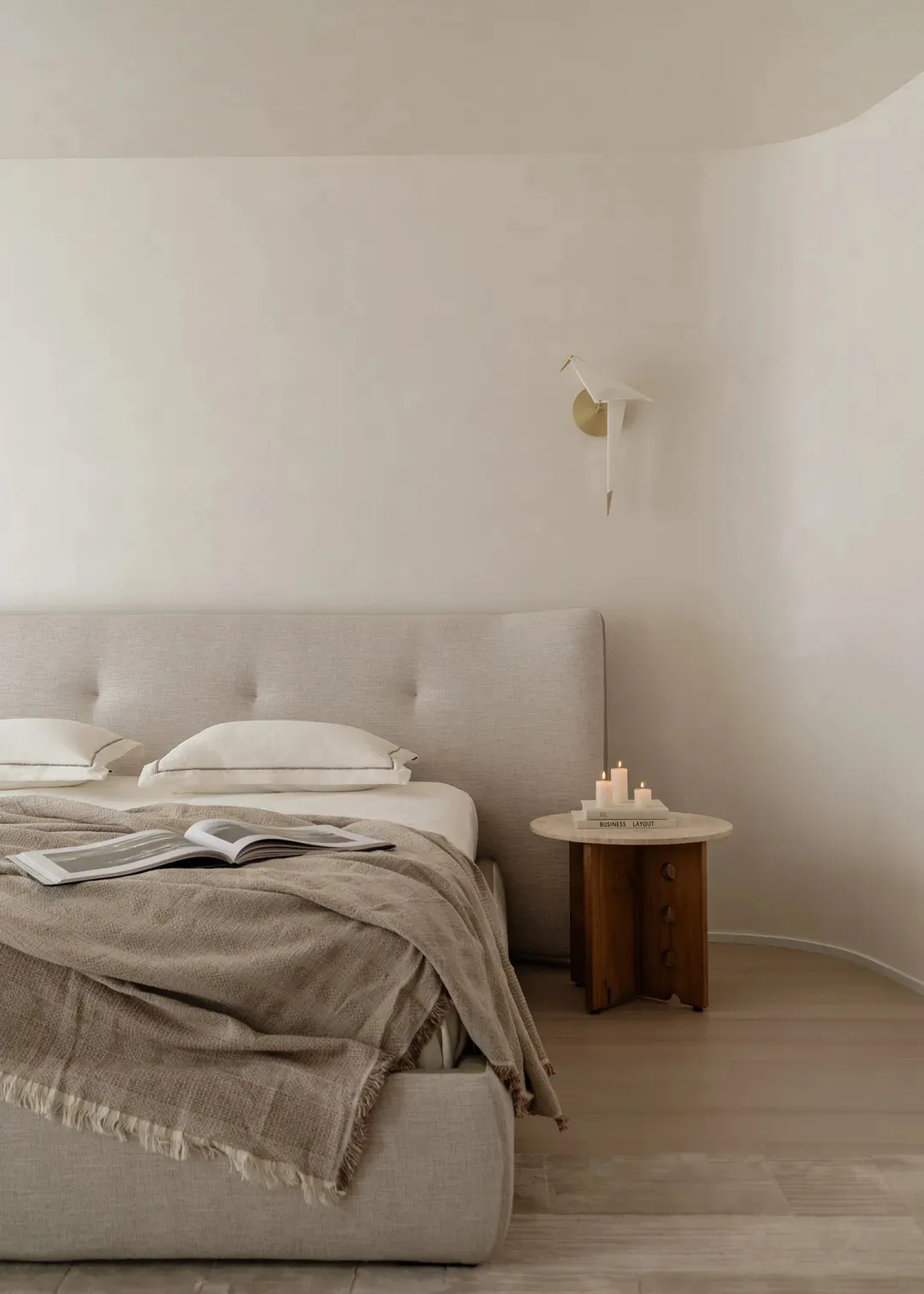 Minimalist bedroom featuring a large upholstered linen bed with beige throw blanket, wooden side table with candles, and a sculptural wall sconce against curved off-white walls.