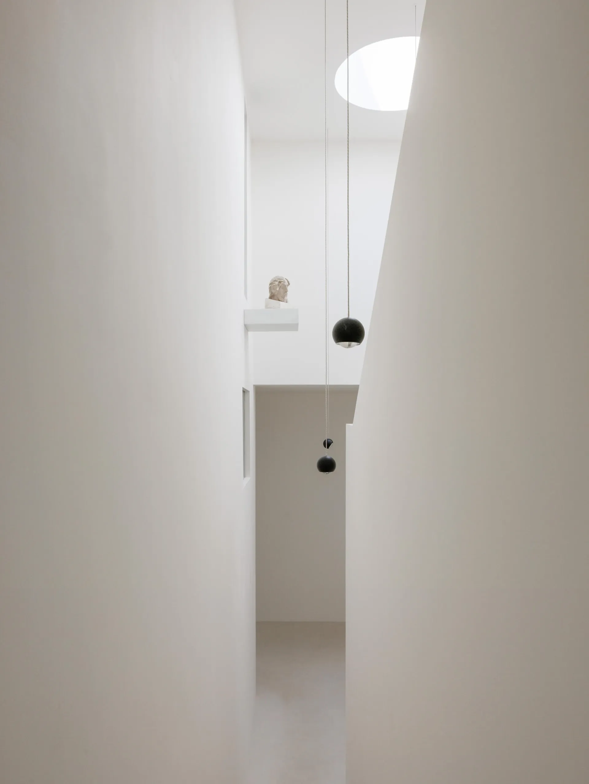 View looking up into a minimalist white double-height hallway featuring two black spherical pendant lights, a classical bust on a high ledge, and a large circular skylight.