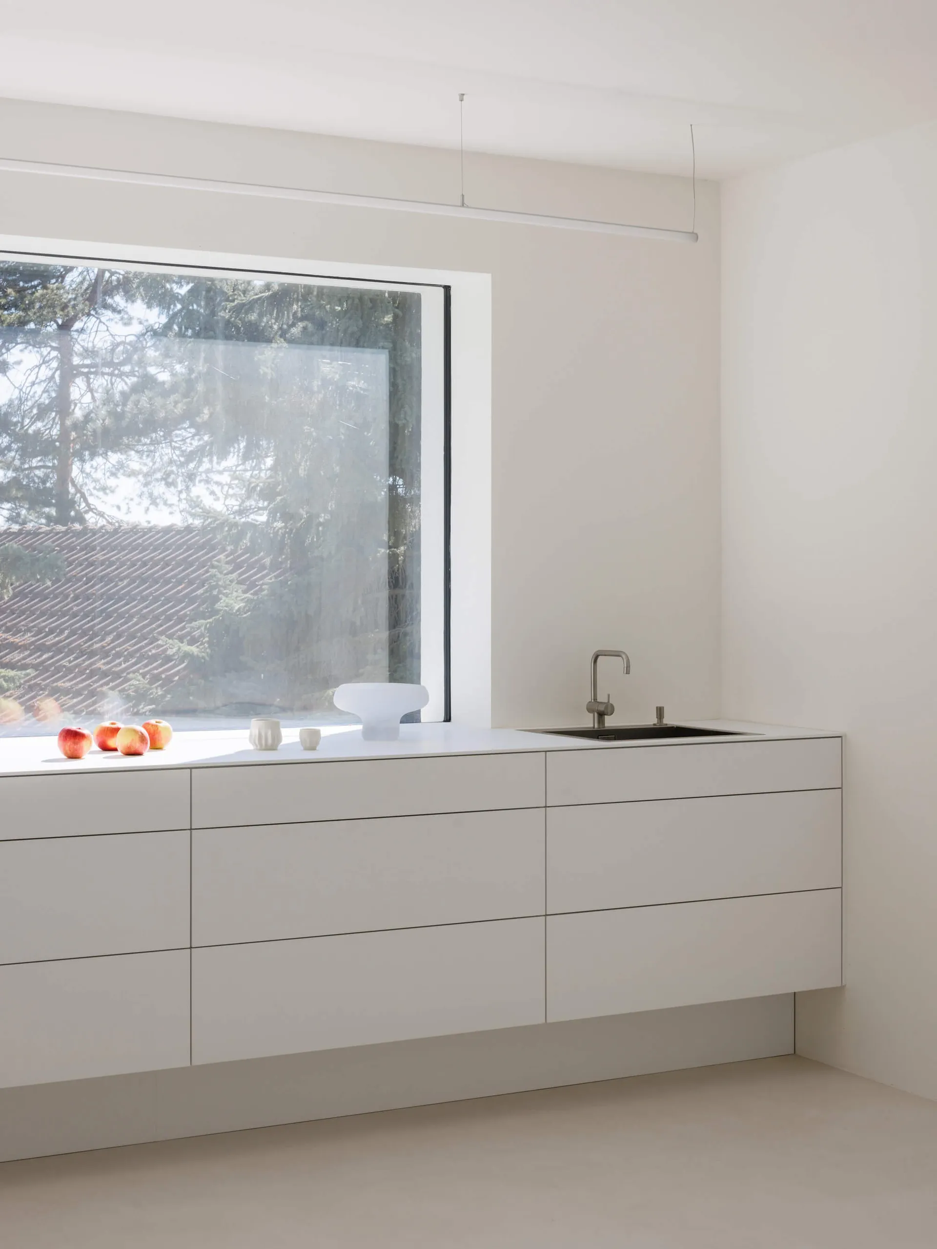 Modern minimalist kitchen featuring handleless white matte lacquer cabinetry, integrated black sink with stainless steel faucet, and a large picture window overlooking trees and a tiled roof.
