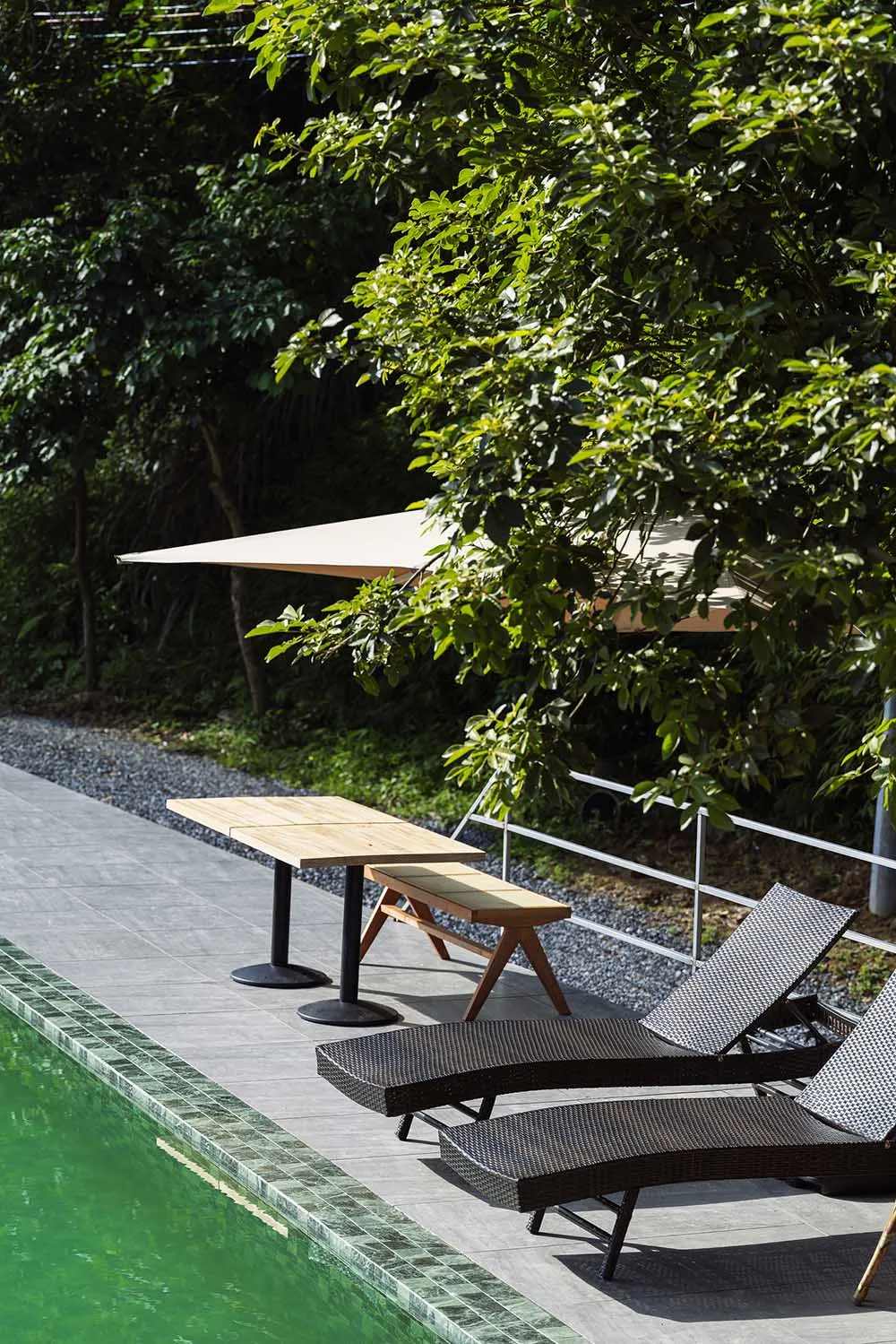 Two dark brown woven wicker lounge chairs on a grey tiled pool deck next to a swimming pool with green water, accompanied by a light wood table and bench under a beige triangular shade sail, surrounded by lush green trees.