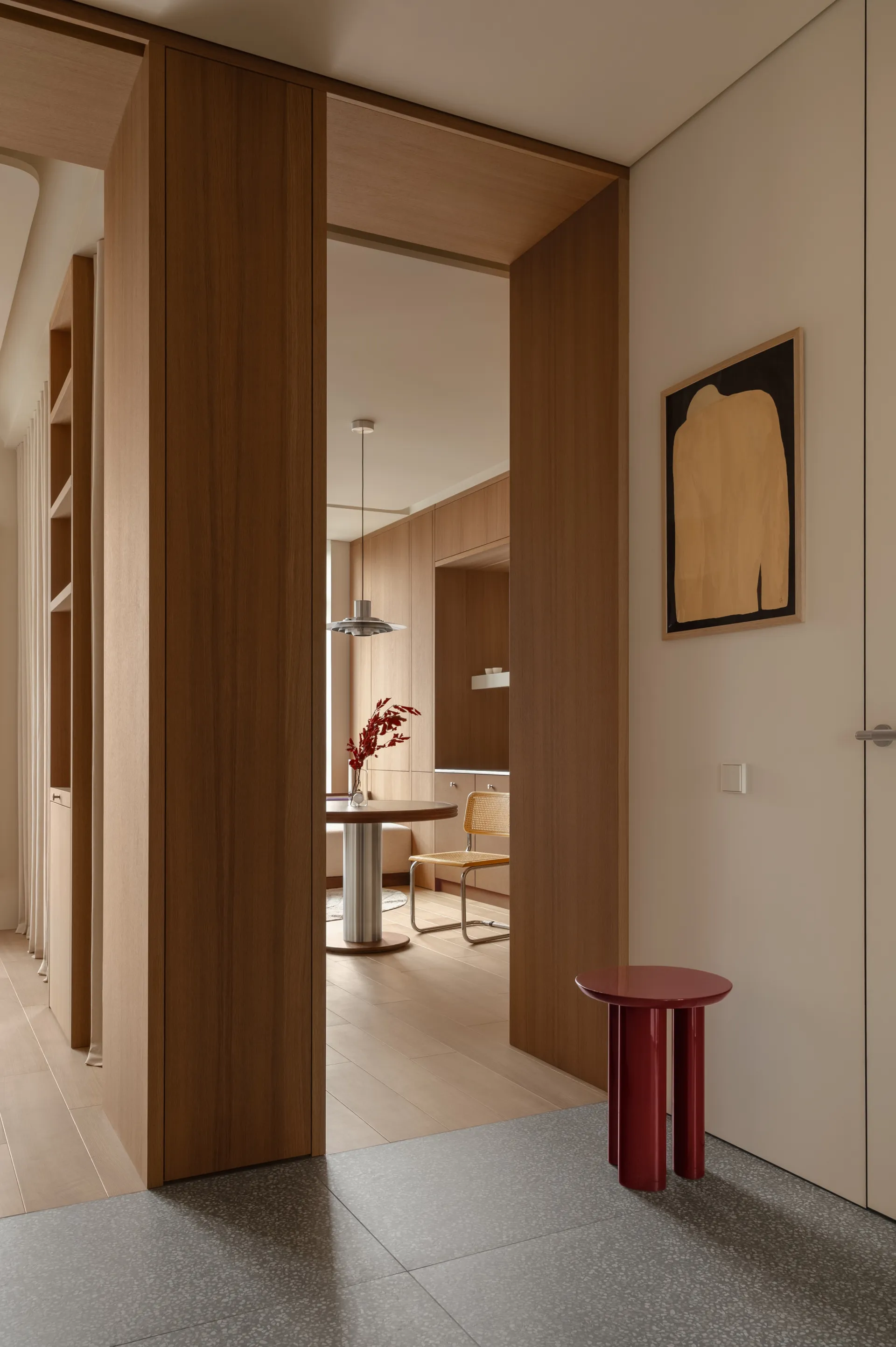 Modern minimalist entryway featuring warm walnut wood cabinetry, a glossy red cylindrical side table, and a view into a light-filled dining area with a round pedestal table and cane chair.