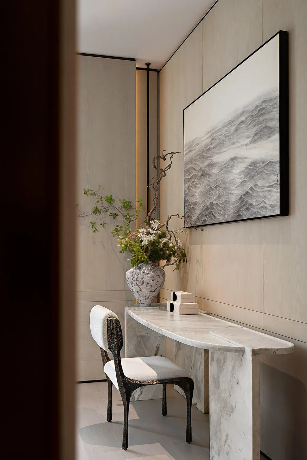 Minimalist entryway featuring a curved white marble console table, a dark sculptural chair, a large ceramic vase with floral arrangement, and a black-framed ink wash painting of waves on a light wood-paneled wall.