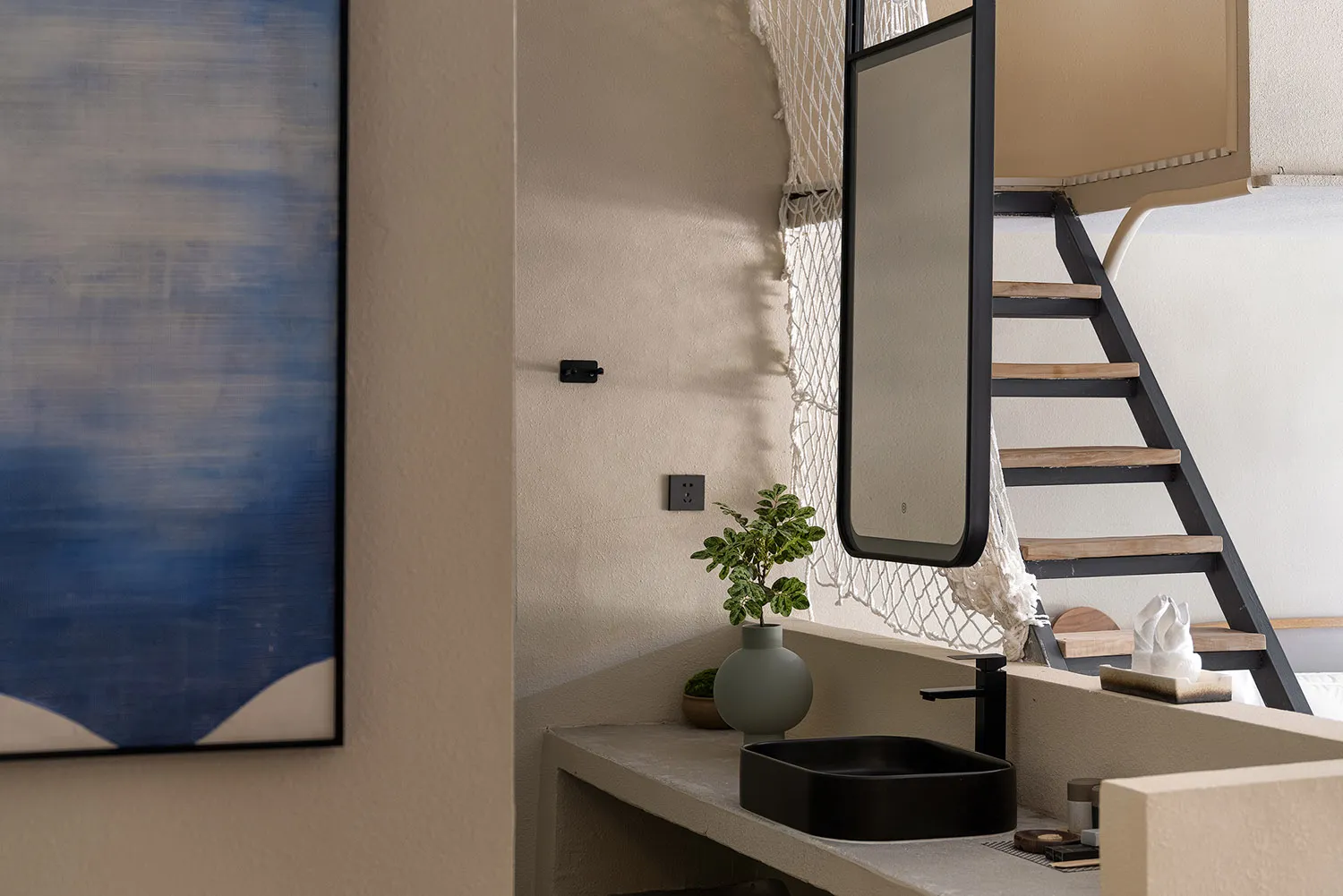 Modern minimalist bathroom vanity made of concrete with a black rectangular vessel sink, black faucet, round mirror, and potted plant against a textured beige wall with a staircase in the background.