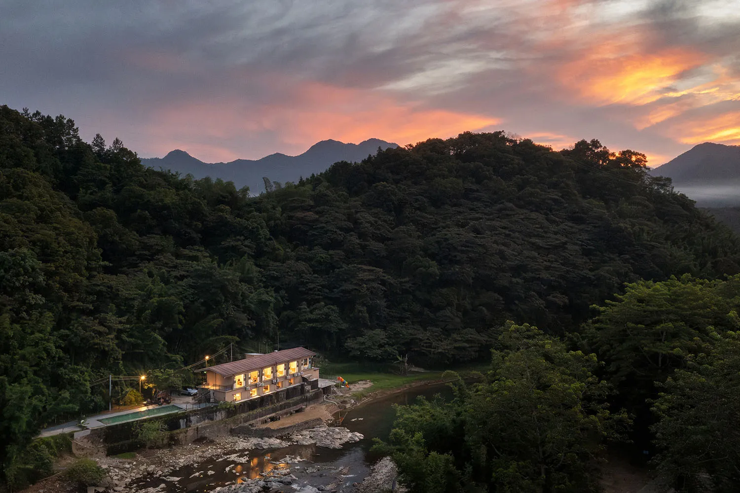 A modern riverside building with warm interior lights glowing at dusk, situated next to a shallow rocky river with forested mountains and a vibrant orange-pink sunset in the background.