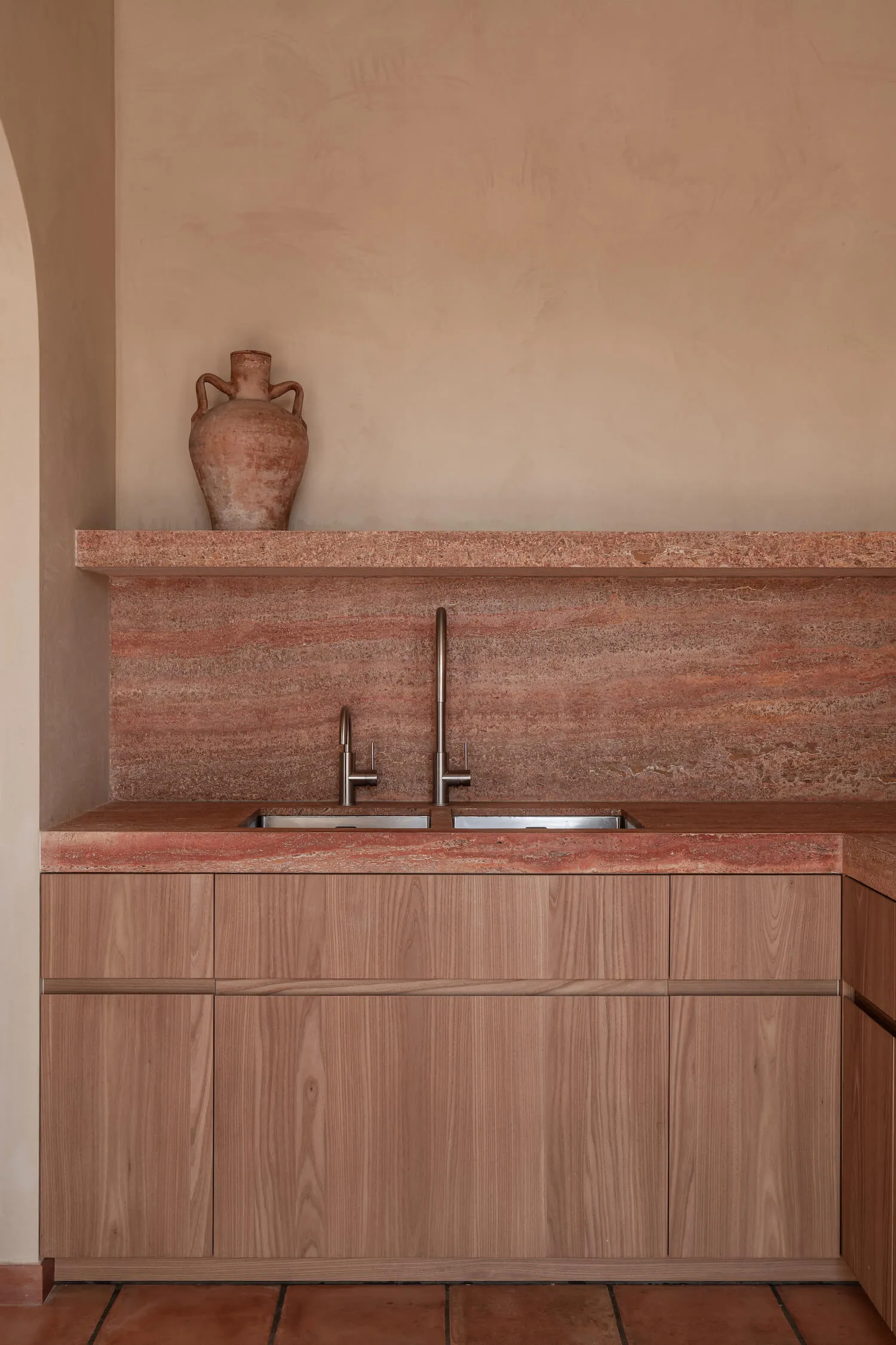 Modern minimalist kitchen featuring a terracotta marble backsplash and countertop, light wood cabinetry, double stainless steel sinks with a tall gooseneck faucet, and a terracotta urn on the shelf against a textured plaster wall.