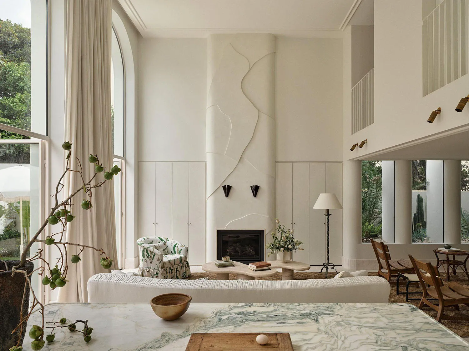 Bright double-height living room featuring a sculptural white plaster fireplace, curved travertine coffee table, white ribbed sofa, and wooden dining chairs with large arched windows overlooking greenery.