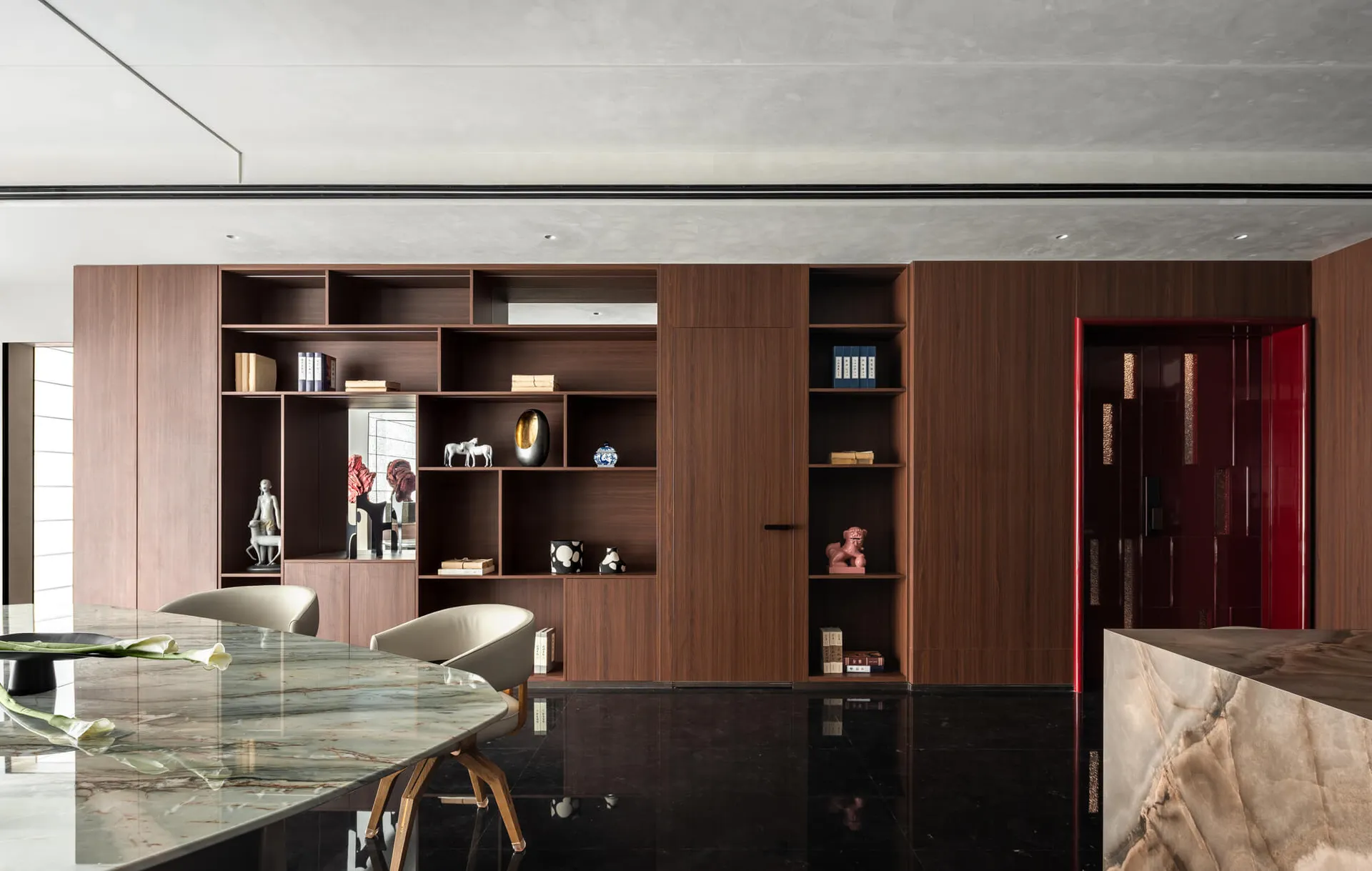 Modern dining room featuring a large walnut built-in shelving unit, marble dining table, cream upholstered chairs, and glossy black floor.