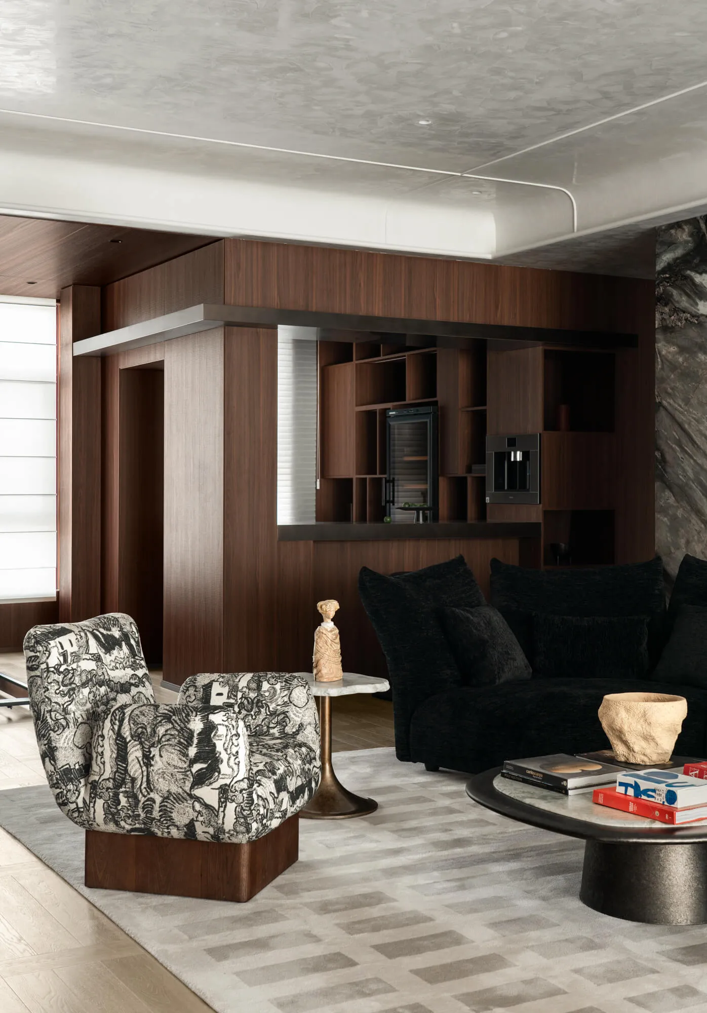 Modern living room with black and white patterned armchair, walnut wood built-in bar, black velvet sofa, marble side table, and grey area rug