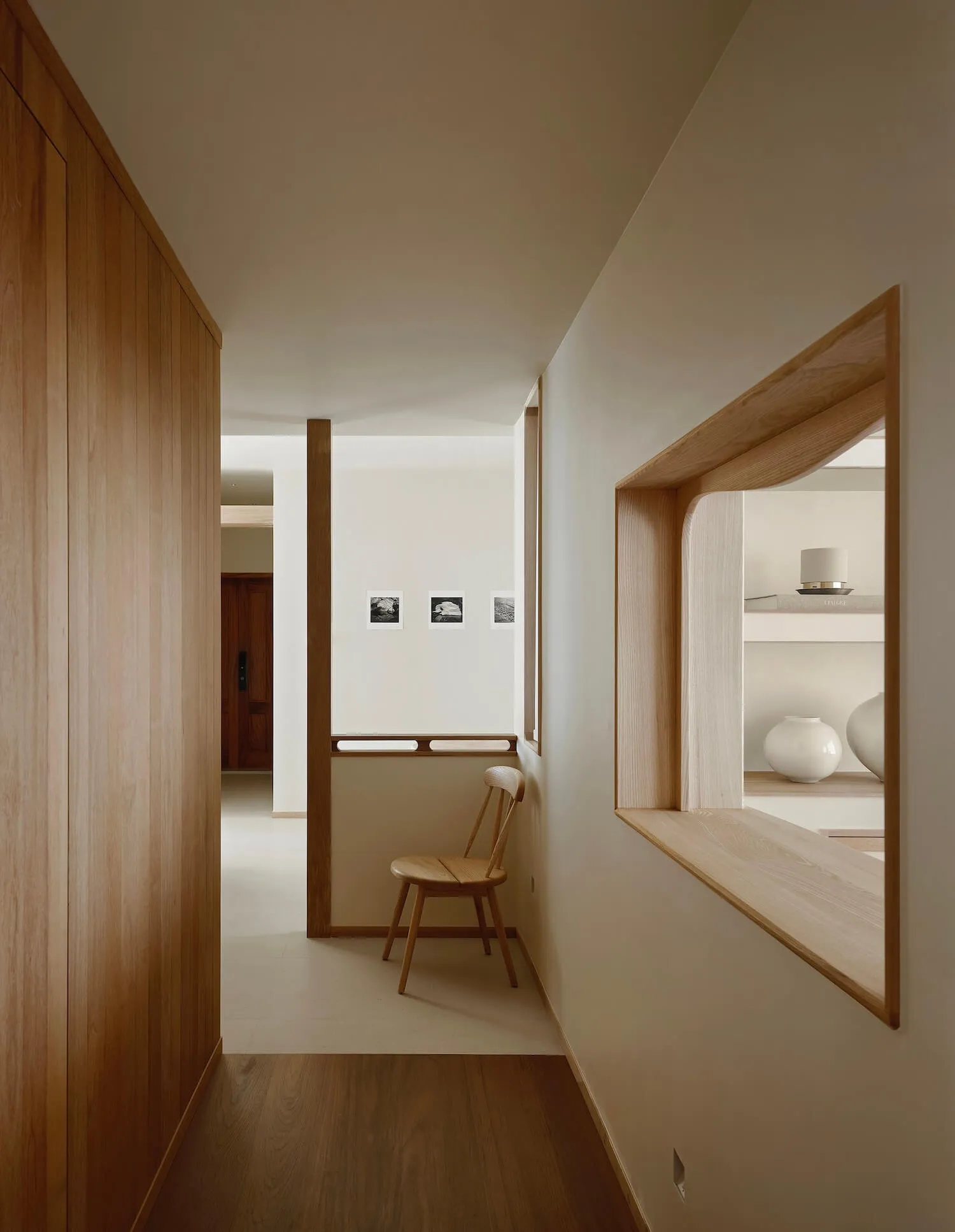 Modern minimalist hallway featuring light oak wood paneling, a built-in curved display niche with white ceramic vases, and a wooden chair against a white wall with small framed photographs.