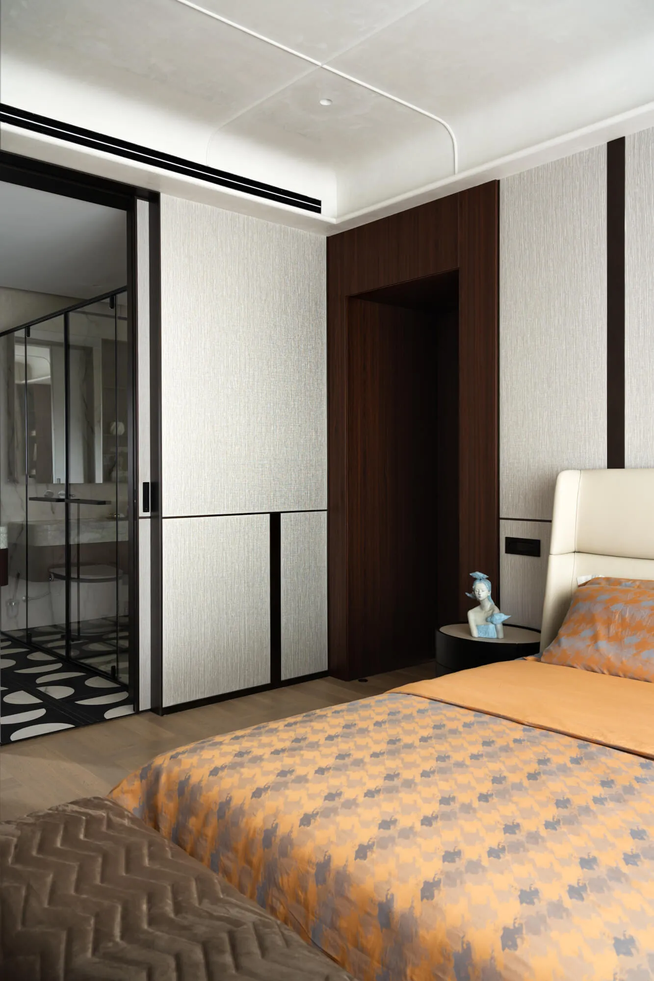Modern luxury bedroom interior with dark walnut wood paneling, light textured walls, orange and grey checker-pattern duvet, cream leather headboard, and black-framed glass door leading to an en-suite bathroom.