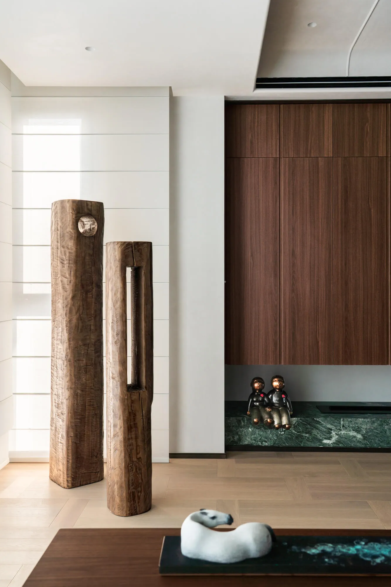 Modern minimalist entryway featuring two large weathered wood sculptures, a walnut wood wall cabinet, green marble ledge with bronze figurines, and a white horse sculpture on a dark console.