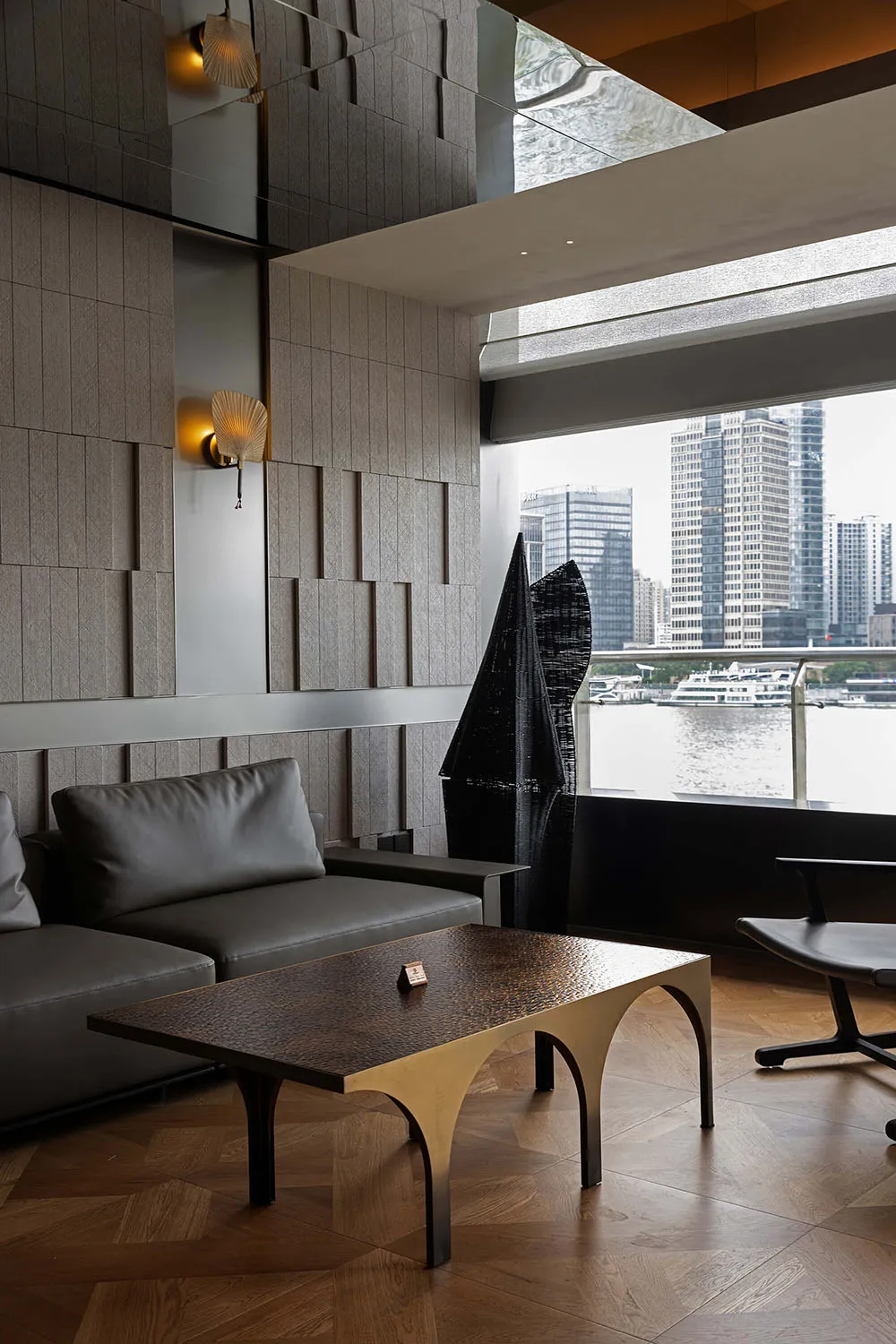 Modern lounge interior featuring a textured dark oak and brass coffee table, grey leather sofa, sculptural black artwork, and large windows overlooking a harbor with city skyline.