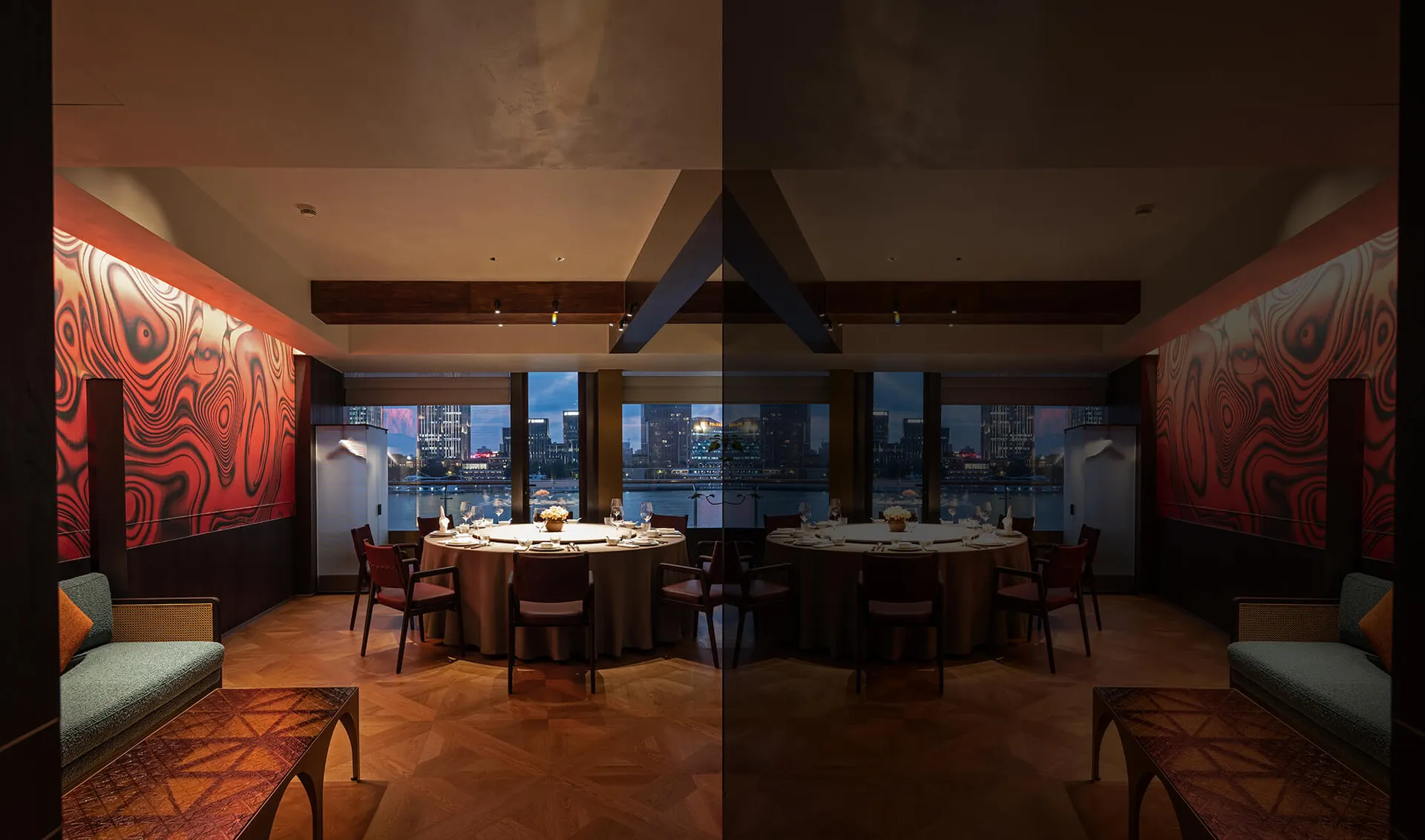 Luxurious private dining room featuring a large round table, red marble accent walls, walnut herringbone flooring, and floor-to-ceiling windows overlooking a harbor city skyline at dusk.