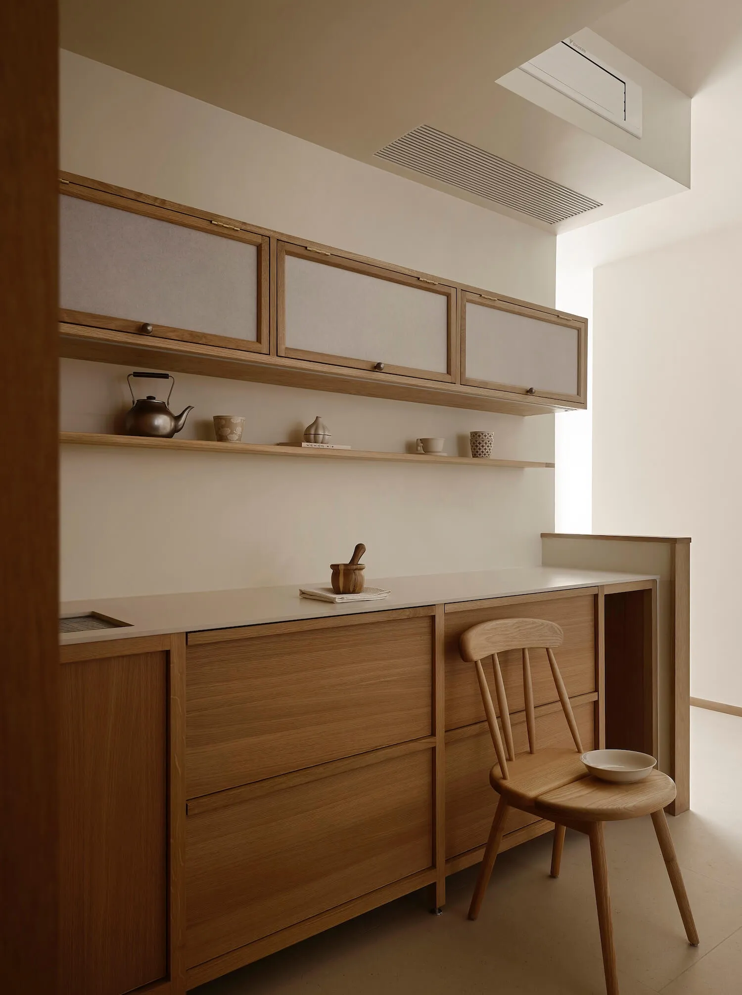 Modern minimalist kitchen with light oak wood cabinetry, white countertops, open floating shelves, wooden chair, and natural light from a skylight.