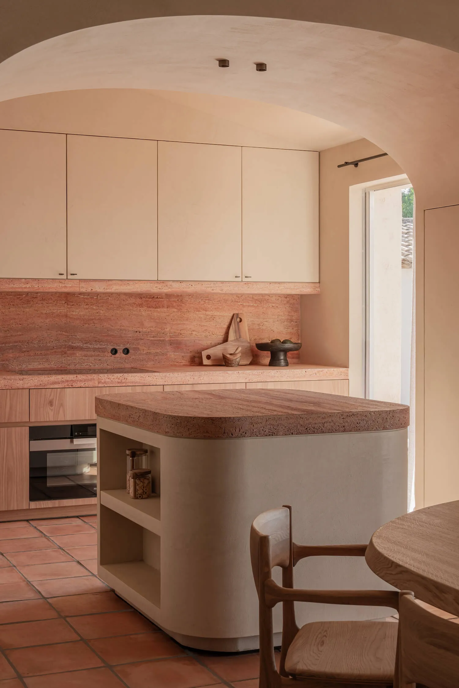 Modern kitchen featuring a curved terracotta kitchen island, matching backsplash, light wood cabinetry, and a large arched plaster ceiling detail with terracotta floor tiles.
