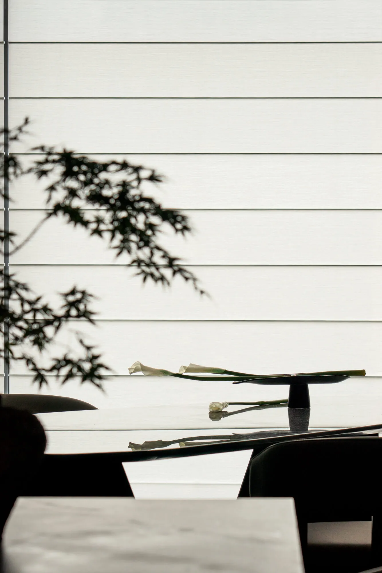 Close-up of a modern glass dining table with black chairs, a sculptural black vase holding white calla lilies, and dramatic shadows of maple leaves cast on a white horizontal-paneled wall.