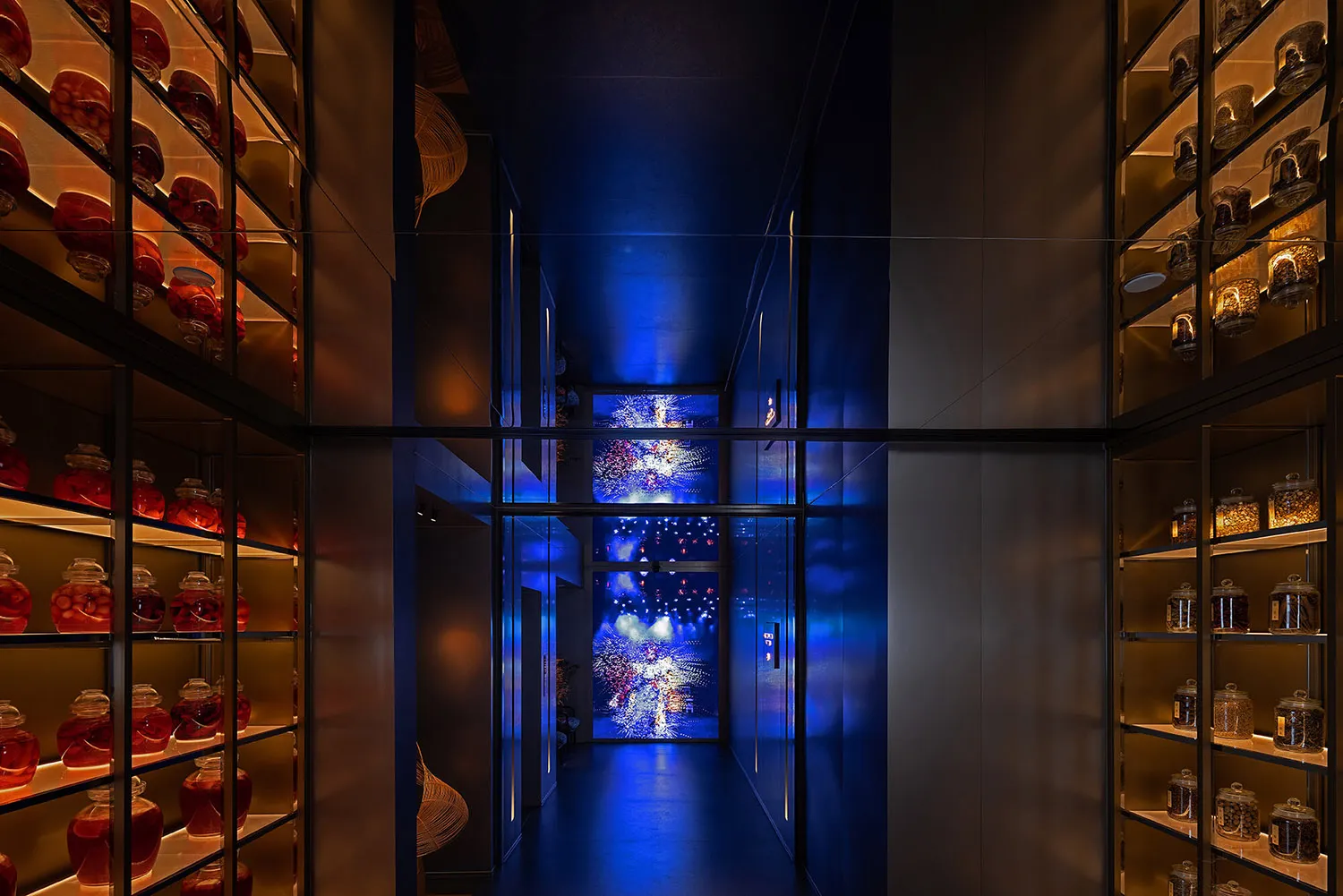 Modern narrow hallway featuring floor-to-ceiling glass and brass shelving filled with illuminated red and amber apothecary jars, dramatic blue ambient lighting, and a large digital firework artwork at the far end.