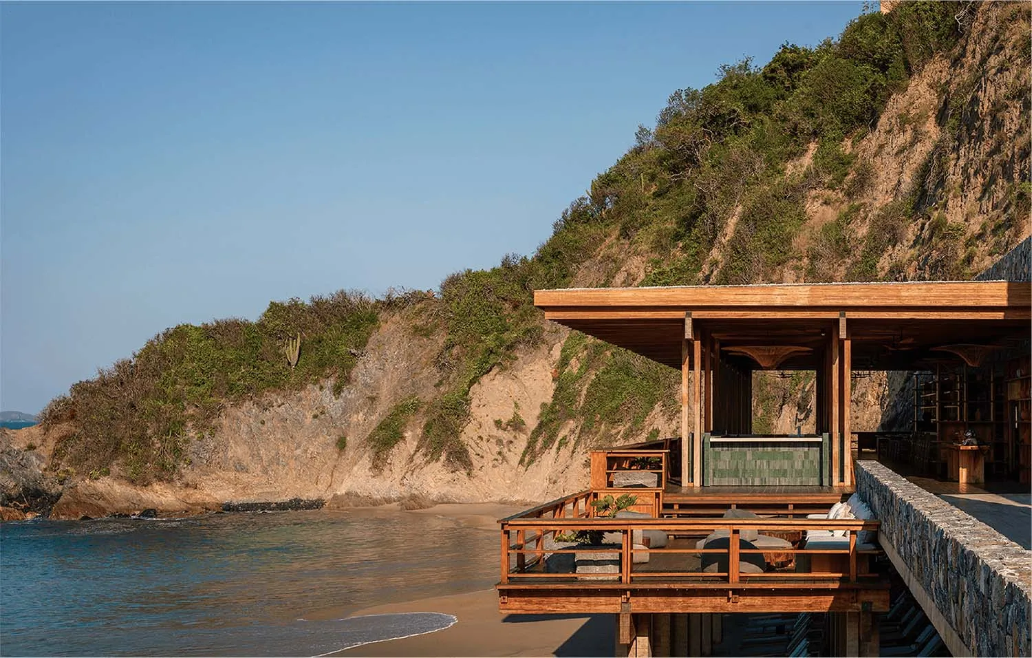 Modern teak wood deck and pavilion built over the ocean at the base of a rocky cliff with lush vegetation under a clear blue sky.