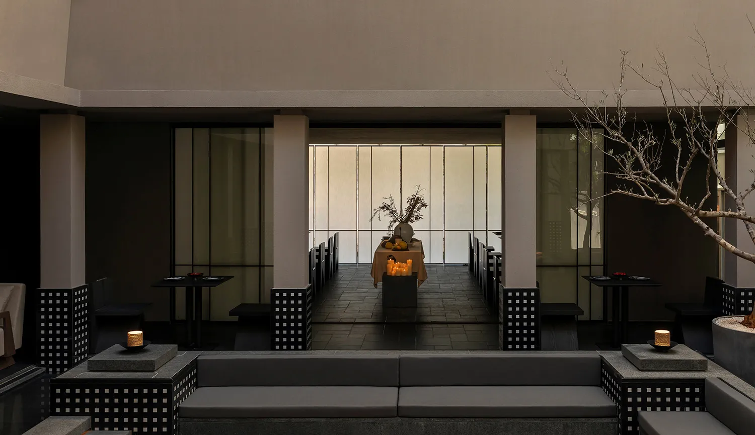 Modern minimalist outdoor dining courtyard at dusk featuring concrete architecture, dark furniture, candlelit tables, and a central dining setup with large windows in the background.