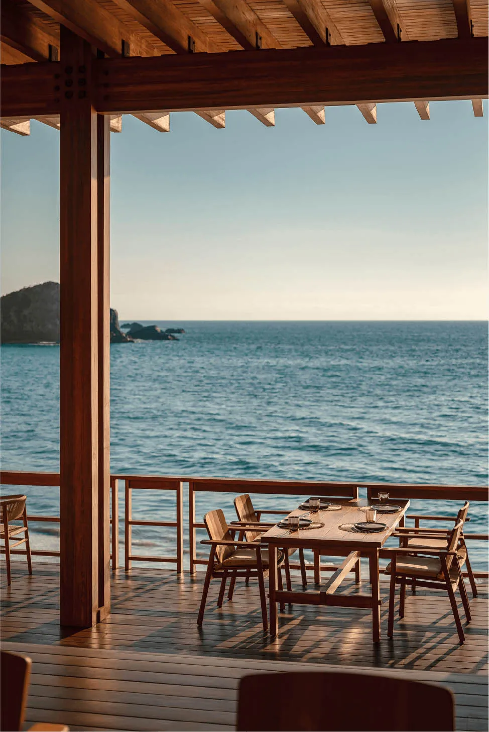 Wooden dining table and chairs on a teak deck under a wooden pavilion overlooking the blue ocean and rocky coastline at sunset