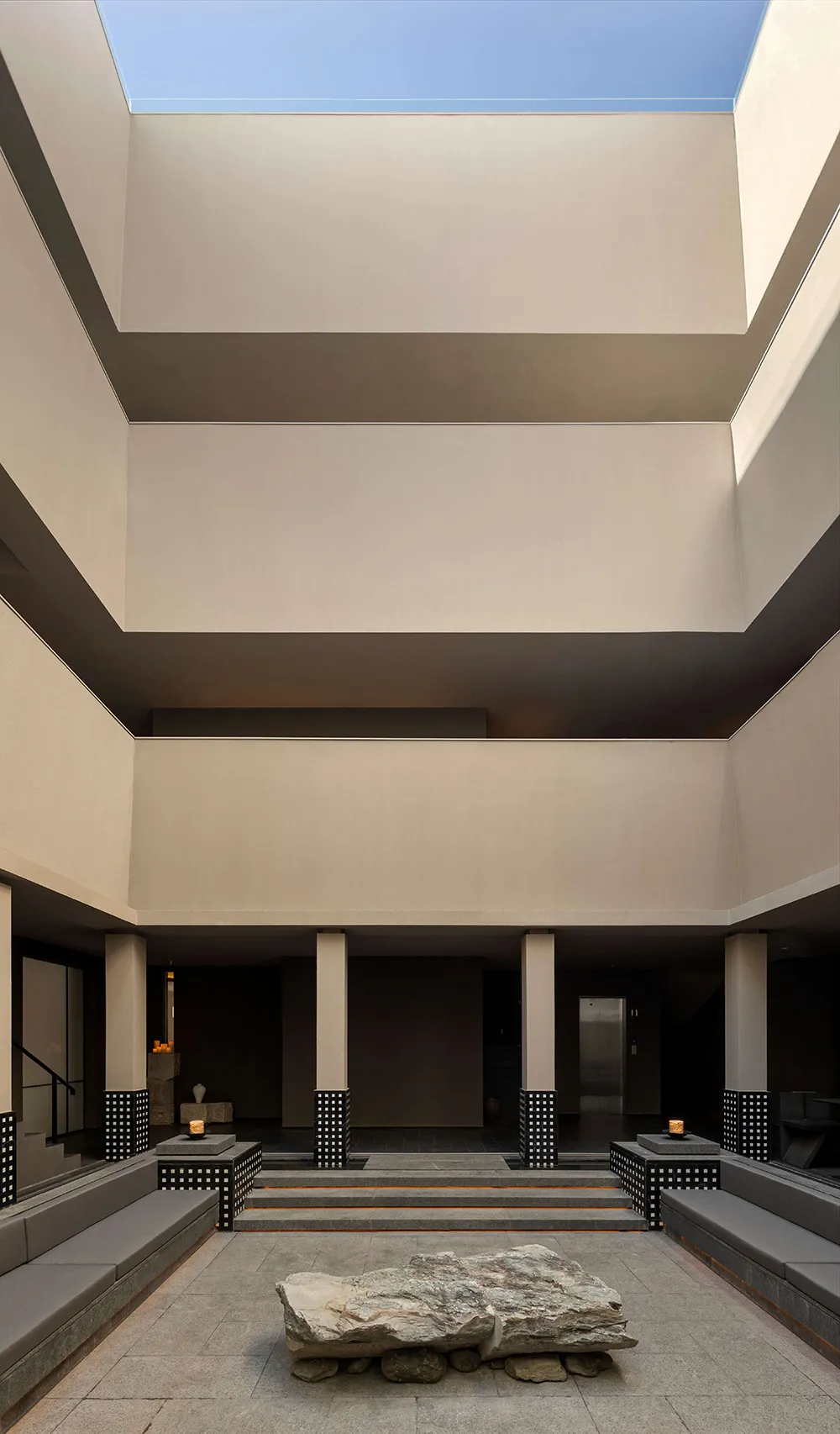 Modern minimalist courtyard with a large natural stone centerpiece, built-in concrete benches, black-and-white patterned columns, and a large rectangular skylight opening to blue sky.