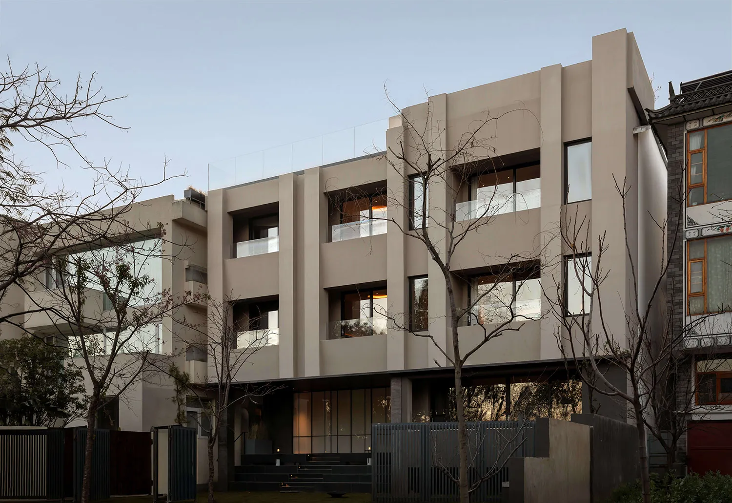 Modern multi-story concrete residential building with large glass windows and cantilevered glass balconies, surrounded by leafless trees at dusk.