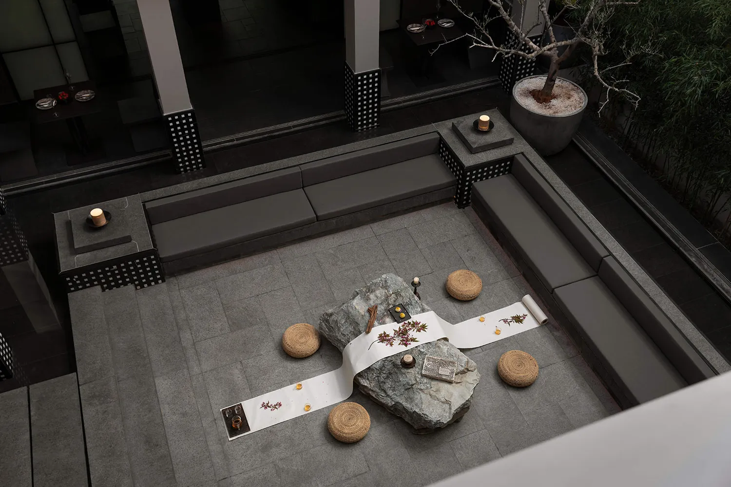 Modern minimalist outdoor courtyard with grey stone pavers, built-in grey benches, large natural rocks, woven straw cushions, and a white embroidered runner set up for a Japanese tea ceremony at night.