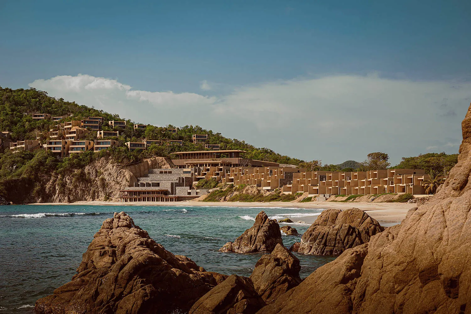 Modern concrete modular resort buildings integrated into a green hillside above a sandy beach and turquoise ocean, viewed from rocky outcrops in the foreground.