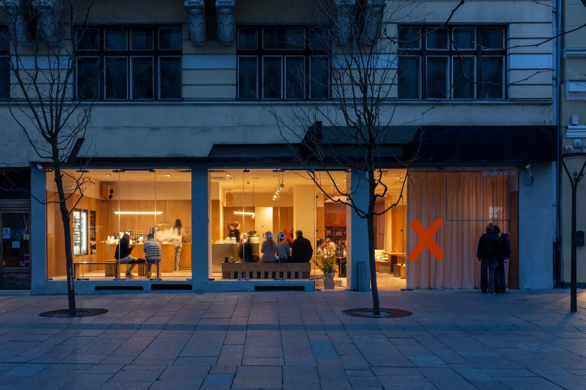 Night view of a modern minimalist cafe named Ukyo X with floor-to-ceiling glass windows revealing warm wooden interiors and patrons inside, located on a paved European street with bare trees.