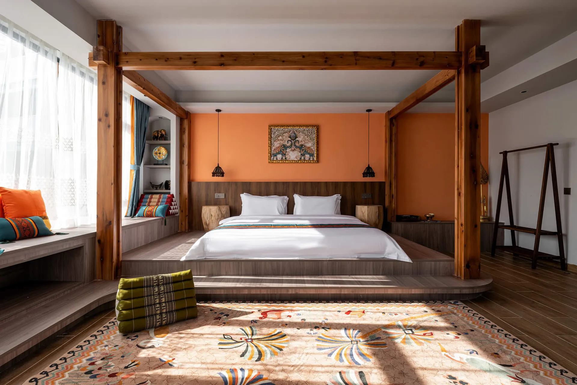 Bright bohemian bedroom featuring a large wooden four-poster bed, burnt orange accent wall, white linens, and colorful patterned rug with sunlight casting strong shadows.