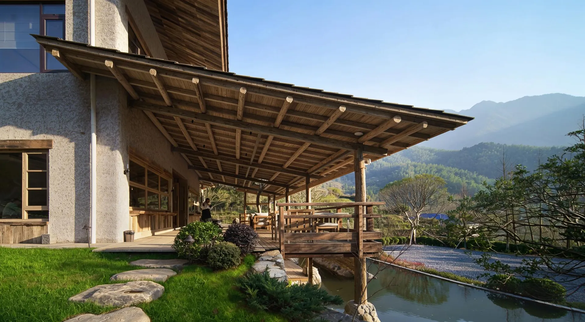 Exterior view of a modern mountain villa featuring a large wooden covered patio, stone stepping path, pond, and distant green mountains under a clear blue sky.