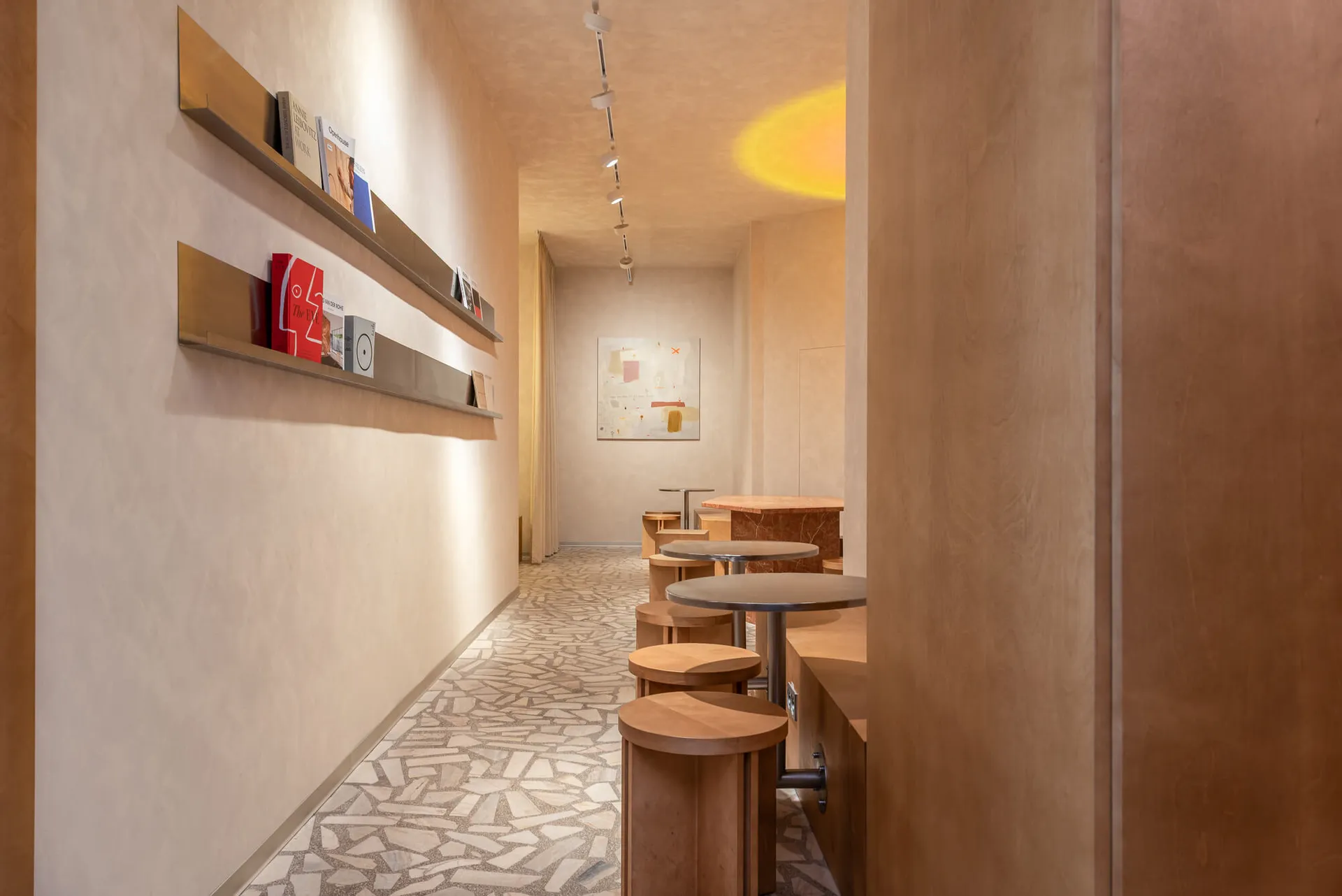 Narrow minimalist cafe interior with terrazzo tile floor, wooden bar stools, brass floating shelves displaying books, and warm ambient lighting.