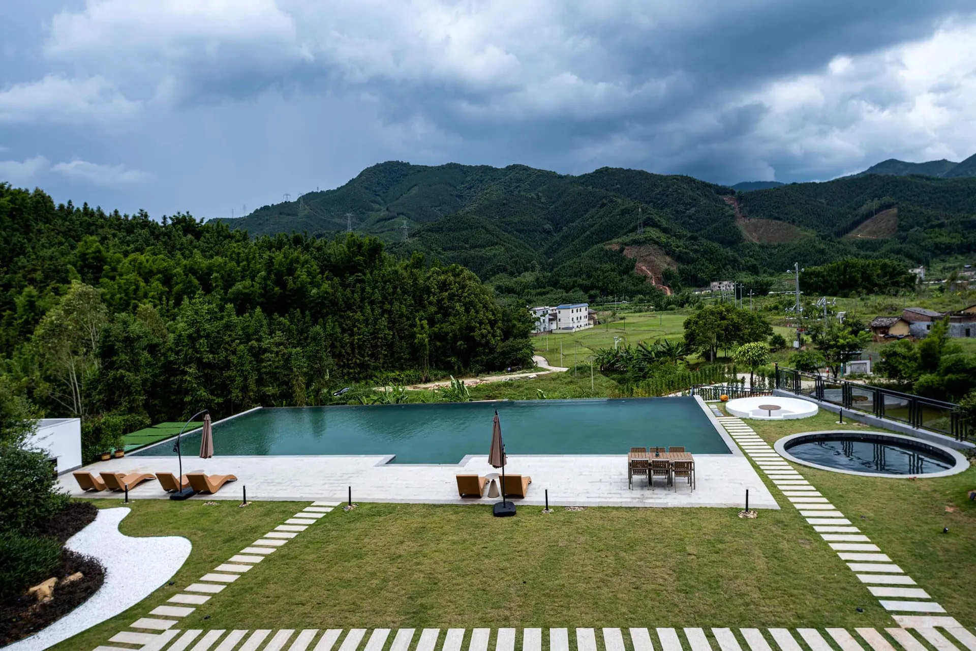 Modern infinity pool and concrete terrace with wooden lounge chairs overlooking lush mountains and rural landscape