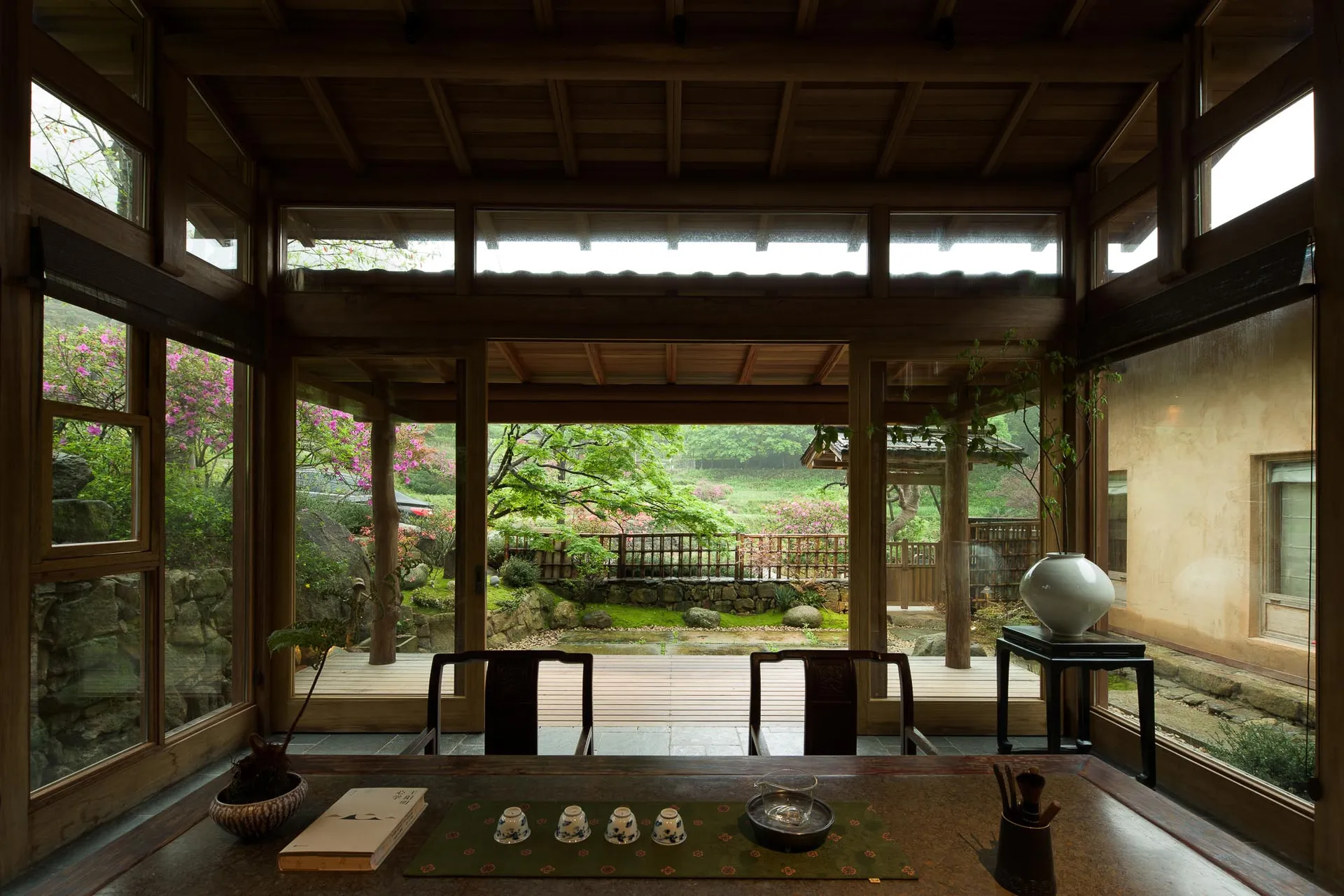 Interior view of a traditional Japanese tea room with dark wooden beams, large glass windows, and a direct view of a lush garden with pink azaleas and a wooden veranda.