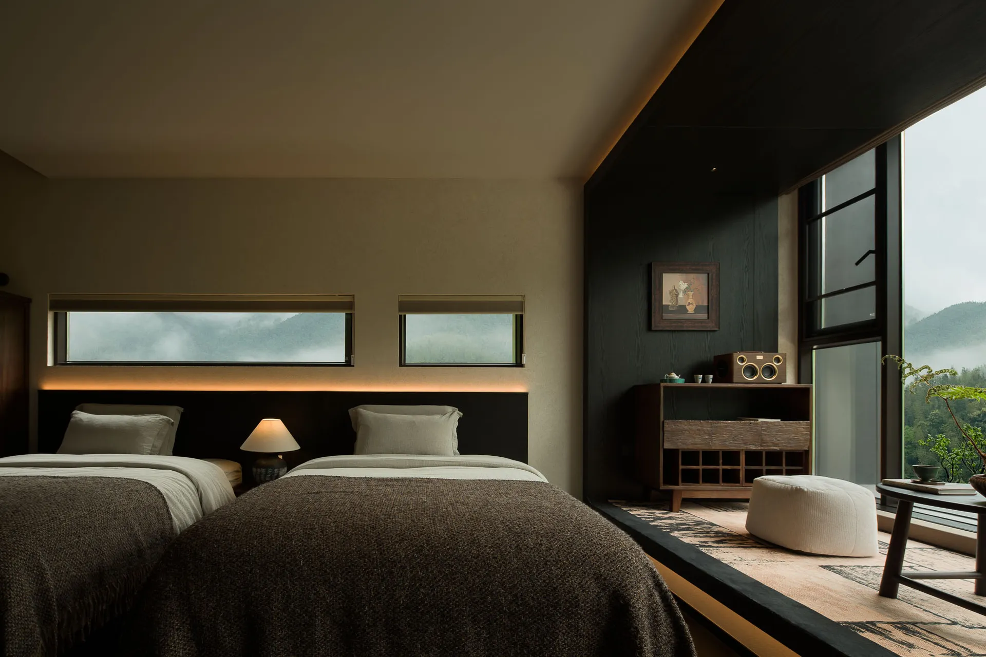 Modern bedroom with two beds covered in brown textured throws, dark wood headboard with backlighting, wooden console with speaker, and large windows revealing a foggy mountain landscape.