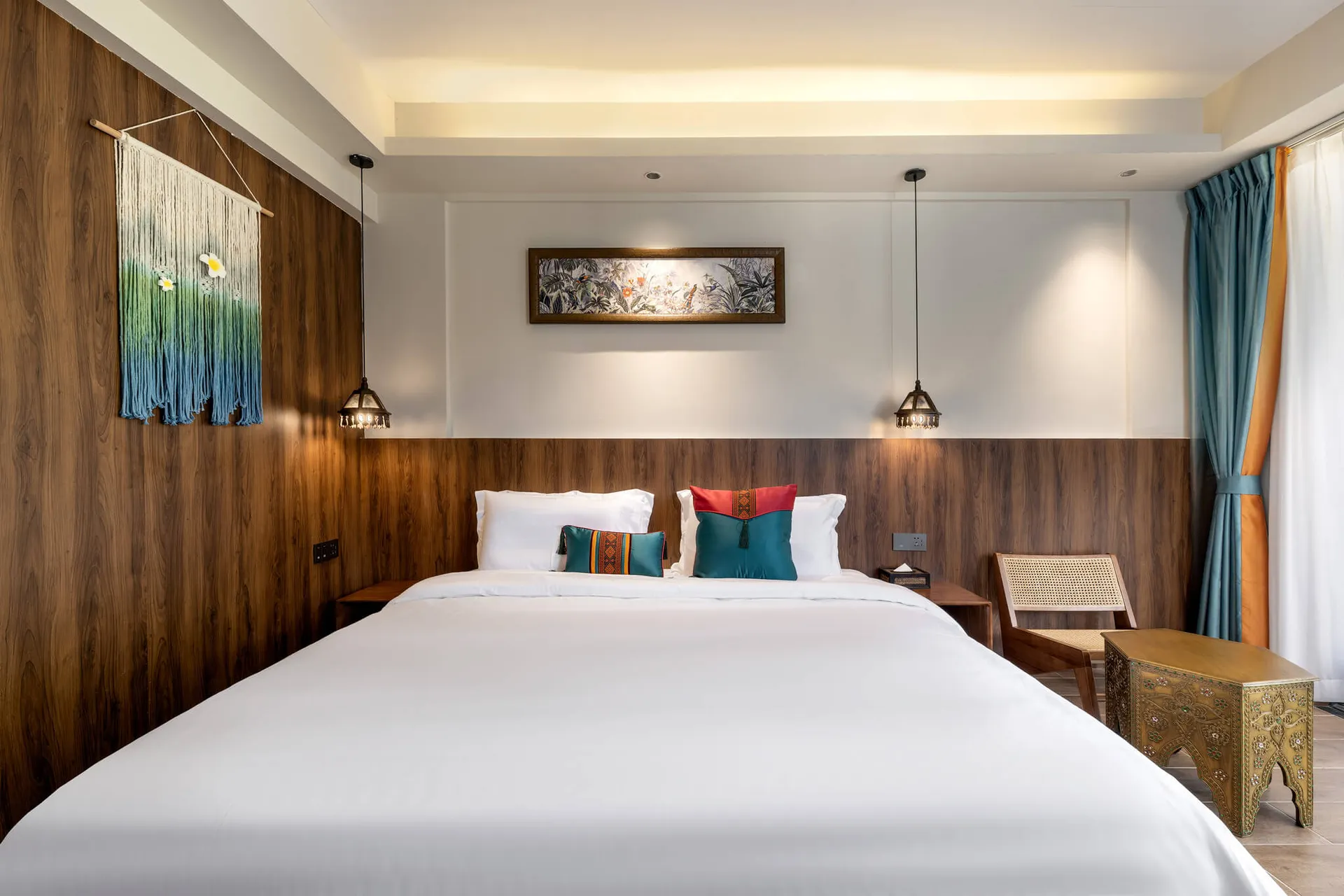 Modern tropical style bedroom featuring a large bed with white linens, colorful teal and red pillows, walnut wood wall paneling, macrame wall hanging, and ornate brass bench.