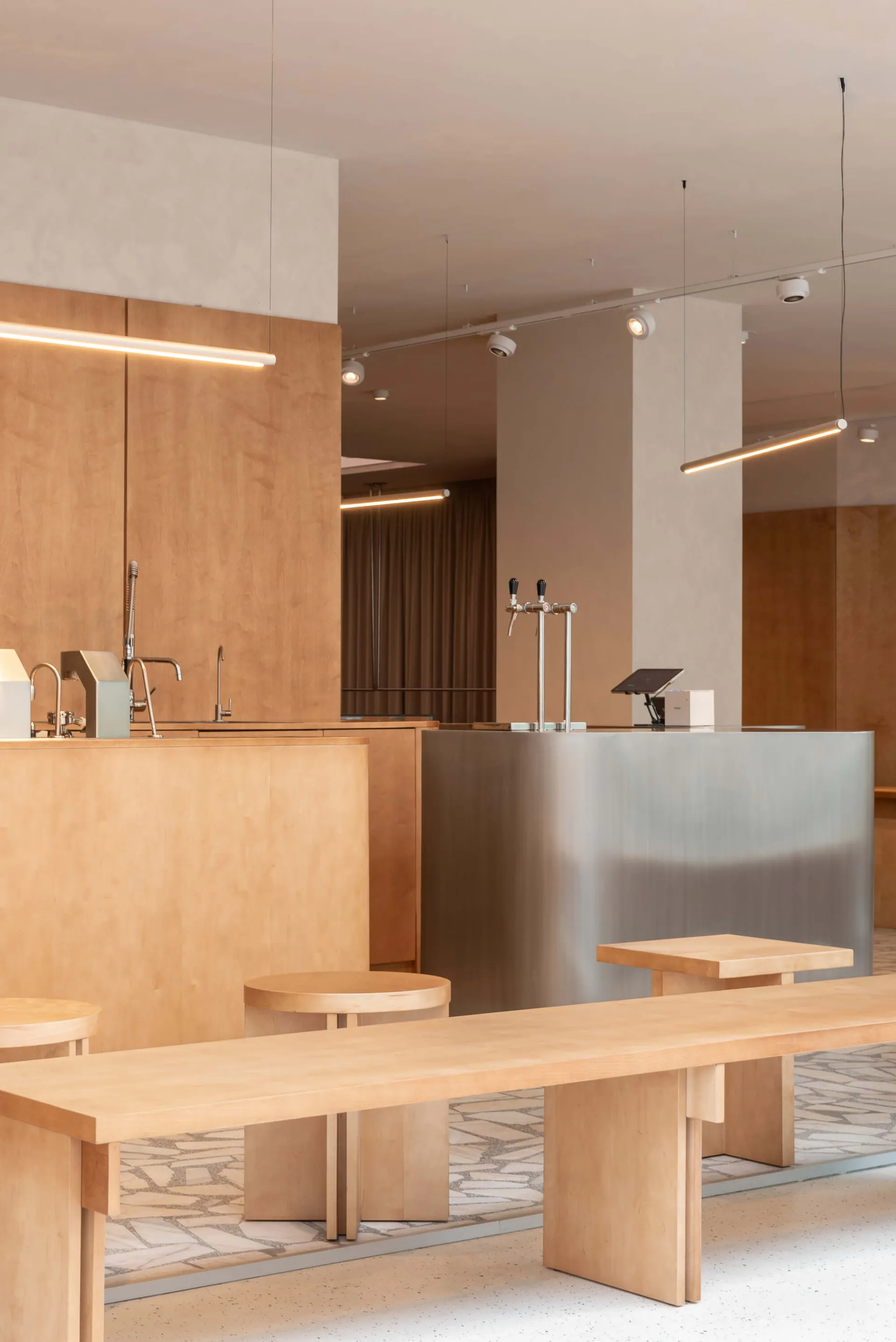 Modern minimalist café interior featuring a long birch plywood communal table, round stools, stainless steel service counter with beer taps, and linear pendant lighting.