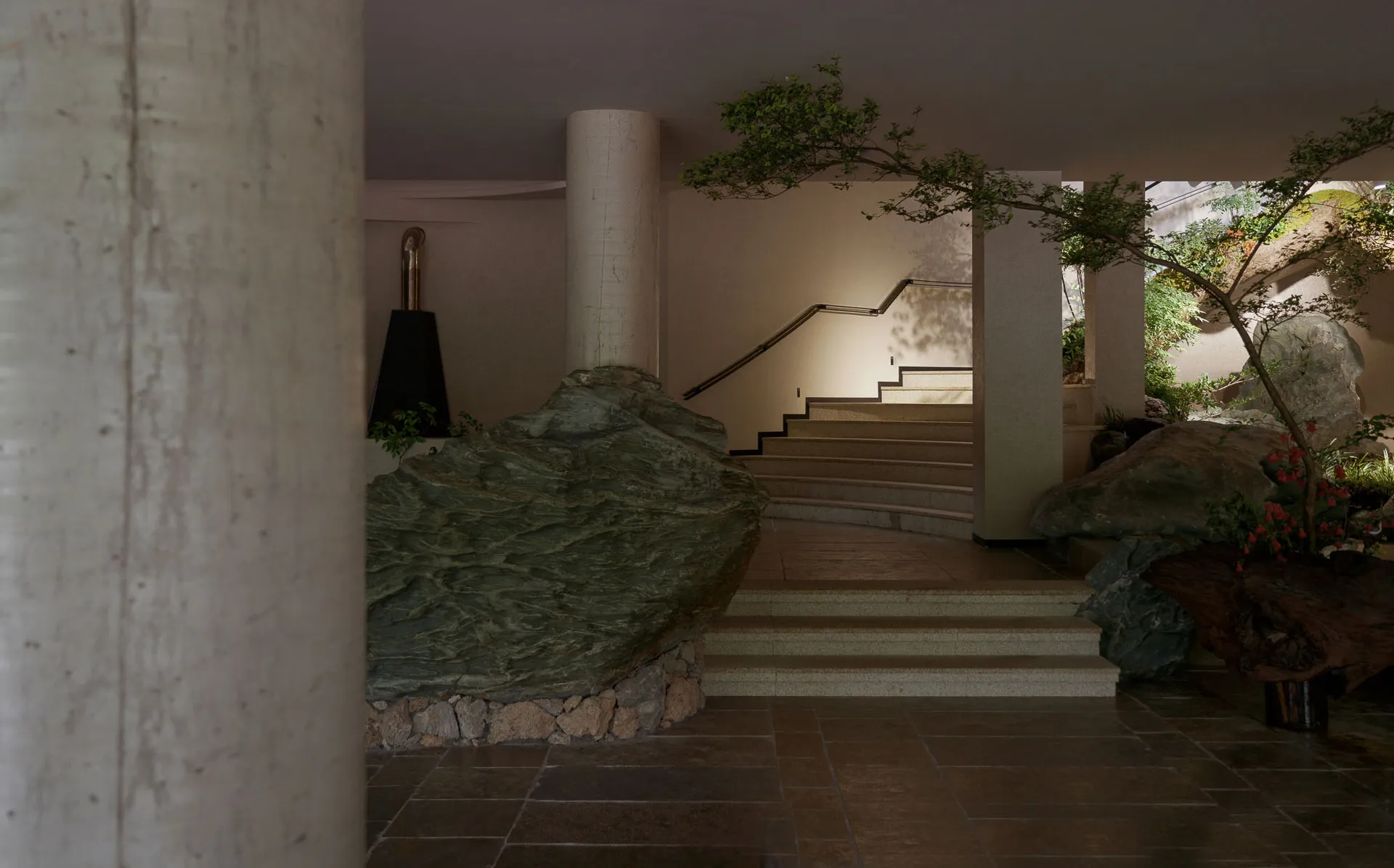 Modern brutalist entrance hall featuring large natural stone boulders, concrete pillars, a curving staircase, and integrated indoor trees under soft ambient lighting.