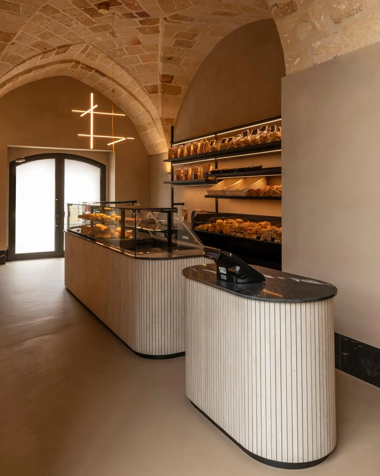 Modern bakery interior featuring a curved fluted oak service counter with black marble top, glass pastry display case, and historic stone vaulted ceiling with modern linear pendant light.
